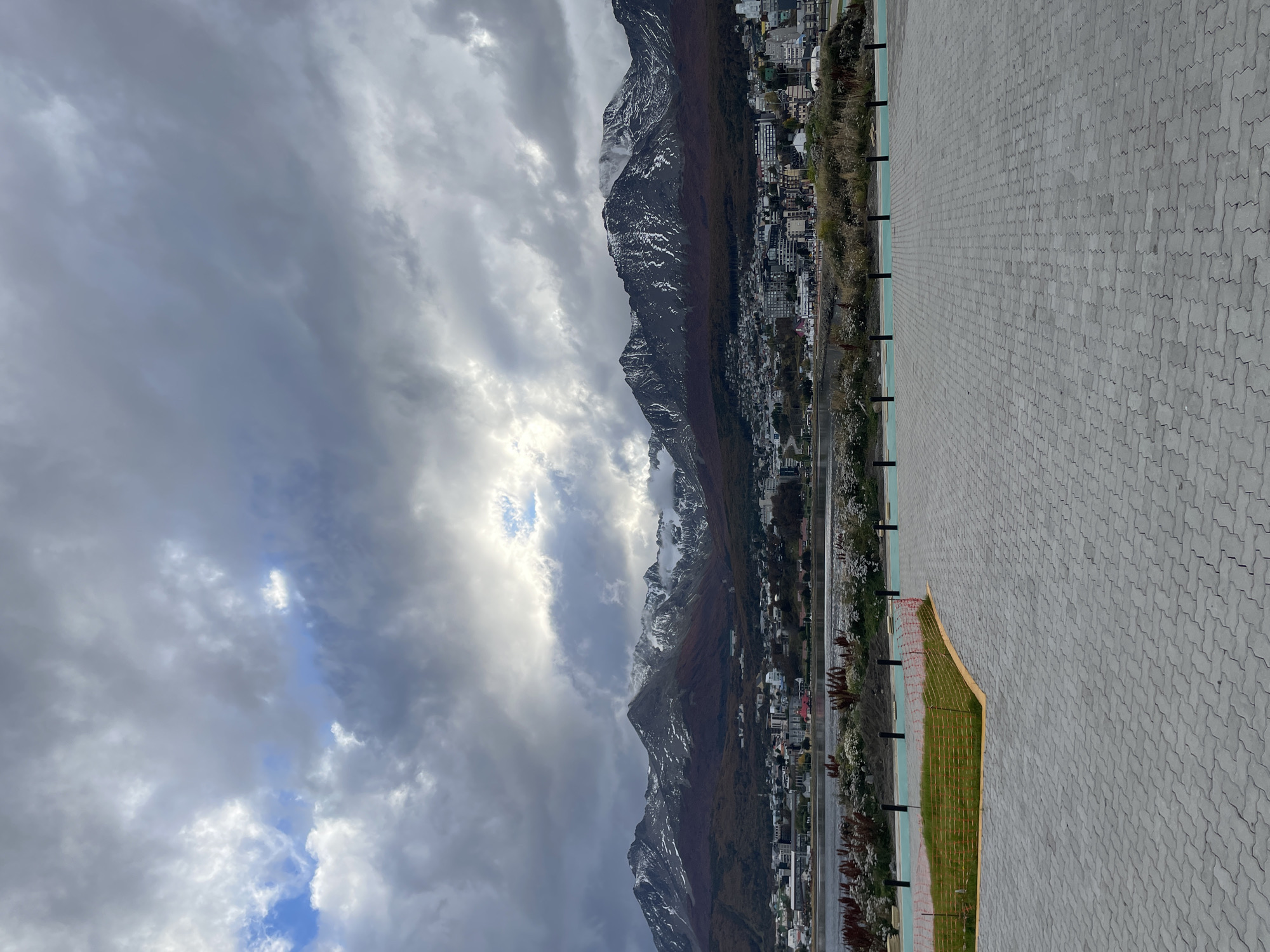 The image captures a mountainous scene in Ushuaia, Argentina, under an overcast sky with glimpses of sunlight breaking through heavy clouds. The landscape features mountains with sparse snow cover, stretching along the edge of a waterfront city. A paved walkway extends into the foreground, marked with black posts. A small patch of green grass is enclosed by yellow mesh netting. The city, nestled at the base of the mountains, is densely built-up with a variety of structures.
