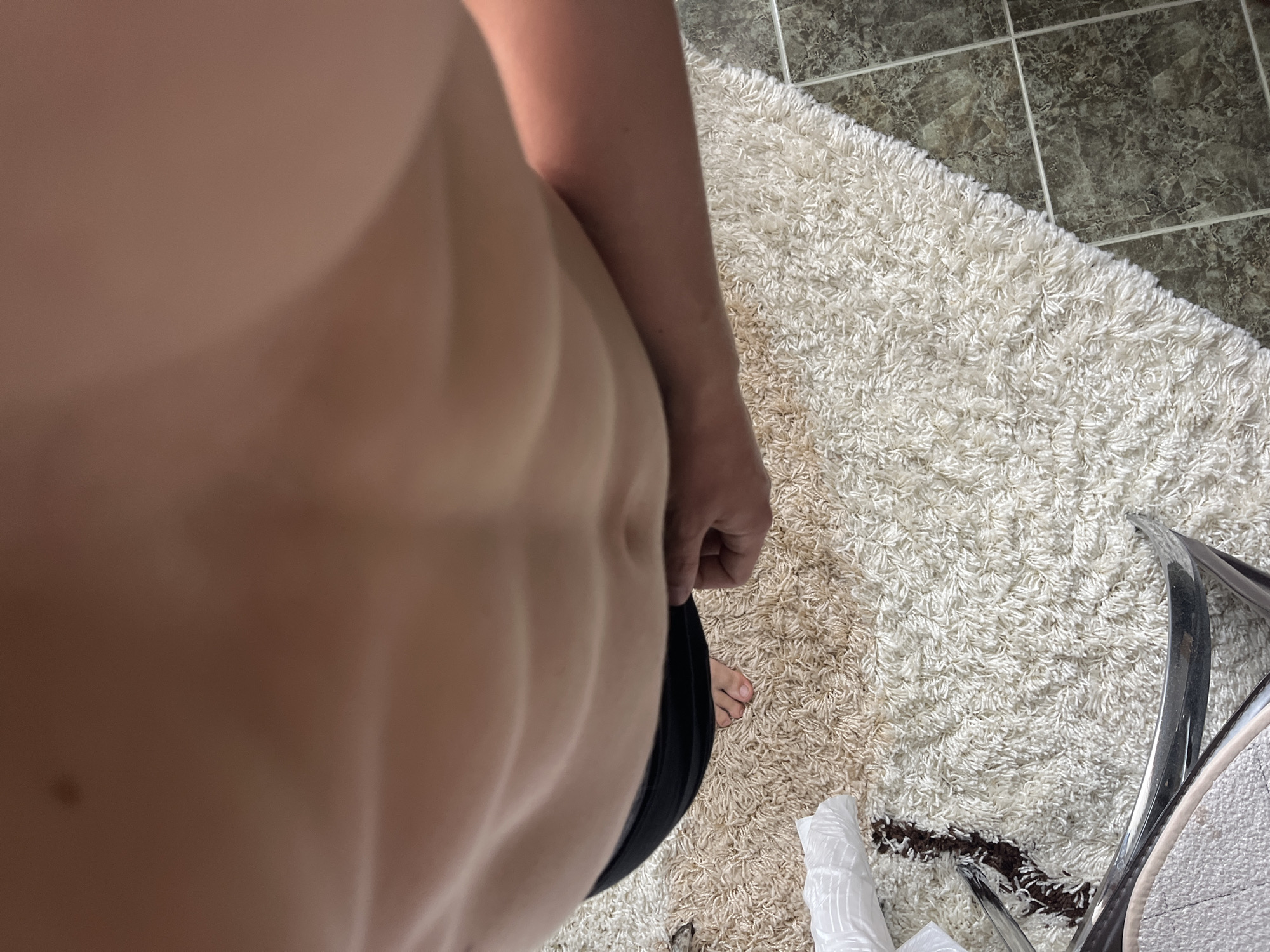 The image captures a downward-facing perspective of an individual's torso and parts of an indoor environment. The central subject is the bare abdomen, displaying well-defined abdominal muscles. A distinct horizontal tan line is visible across the upper abdomen. The individual has fair skin. Part of a left arm is visible along the left edge of the frame, and a hand is partially visible near the center-right, appearing to hold or adjust the black waistband of a lower garment, which could be shorts or underwear. A bare right foot with painted toenails (pink/red) is visible on the rug in the lower right quadrant.
The background consists mainly of a shaggy, light-colored (off-white/cream) rug or carpet that covers most of the right and lower portions of the scene. Along the upper-left, a section of dark, mottled floor tiles with visible grout lines is present, suggesting an adjacent tiled area. In the upper-right, a curved, reflective metallic object, likely a furniture leg or frame, extends into the frame. A crumpled white object (possibly paper or fabric) is visible on the rug near the bottom-right, adjacent to the metallic item. Some darker, possibly soiled or worn, patches are visible on the rug. The lighting suggests an indoor setting.
While the prompt requests the location to be in the city of Zlatograd, Bulgaria, there is no discernible visual information within the image itself to confirm this specific geographic location.