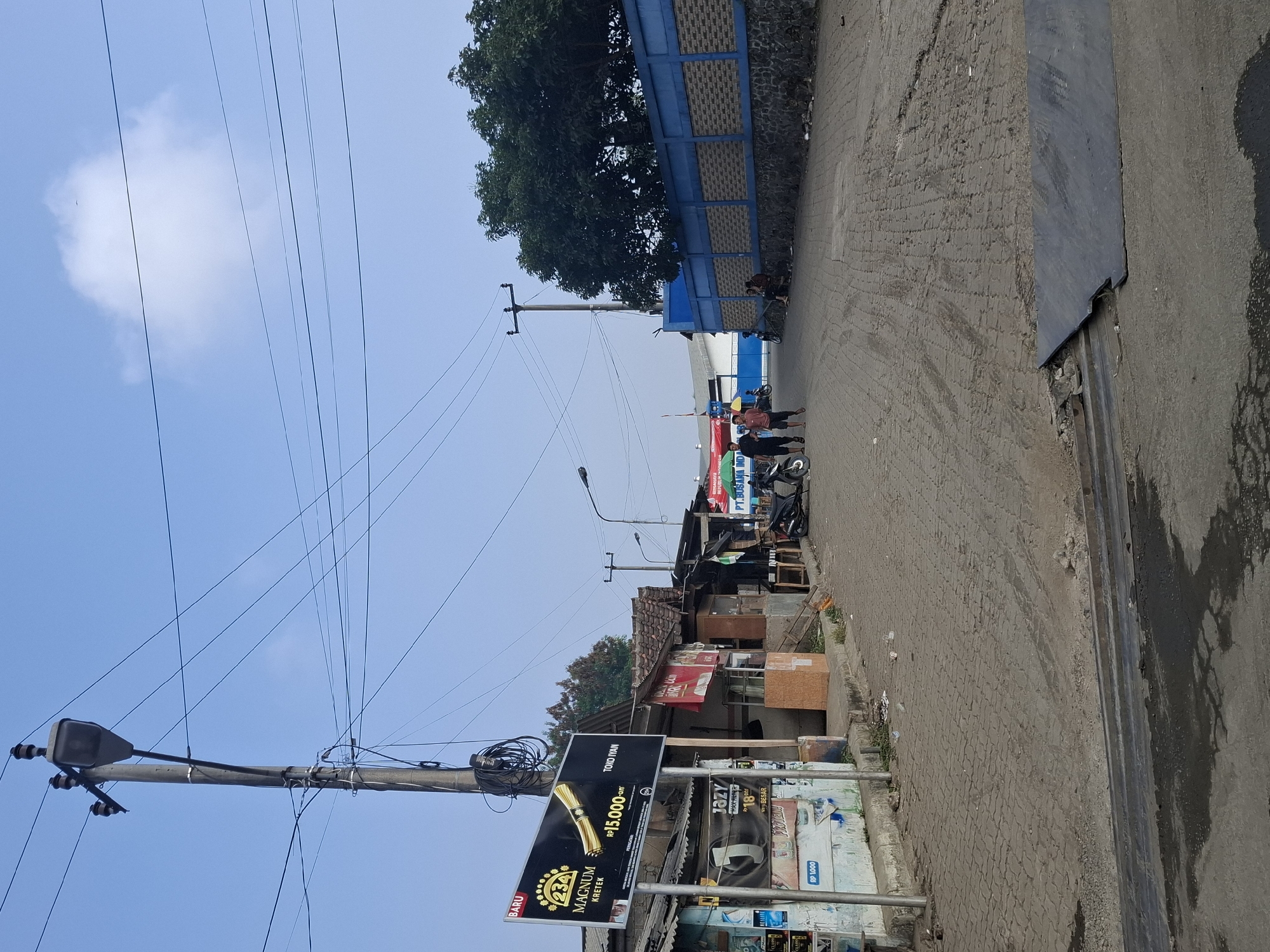 The image depicts a street scene in Ciheulang Tonggoh, Indonesia. A concrete and paver block road runs through the center of the frame, with a sidewalk and various establishments lining the left side. A prominent billboard advertising "MAGNUM KRETEK" is mounted on a utility pole on the left. Several small shops with corrugated metal roofs and open fronts are visible. On the right side of the street, a blue and grey brick wall borders a fenced area with trees behind it. Two people are walking on the road in the mid-ground, and a group of three people are seated on the wall on the right. A motorcycle is parked in front of one of the shops. The sky is blue with a few scattered clouds. The overall environment appears to be a commercial or mixed-use area with a somewhat weathered appearance.