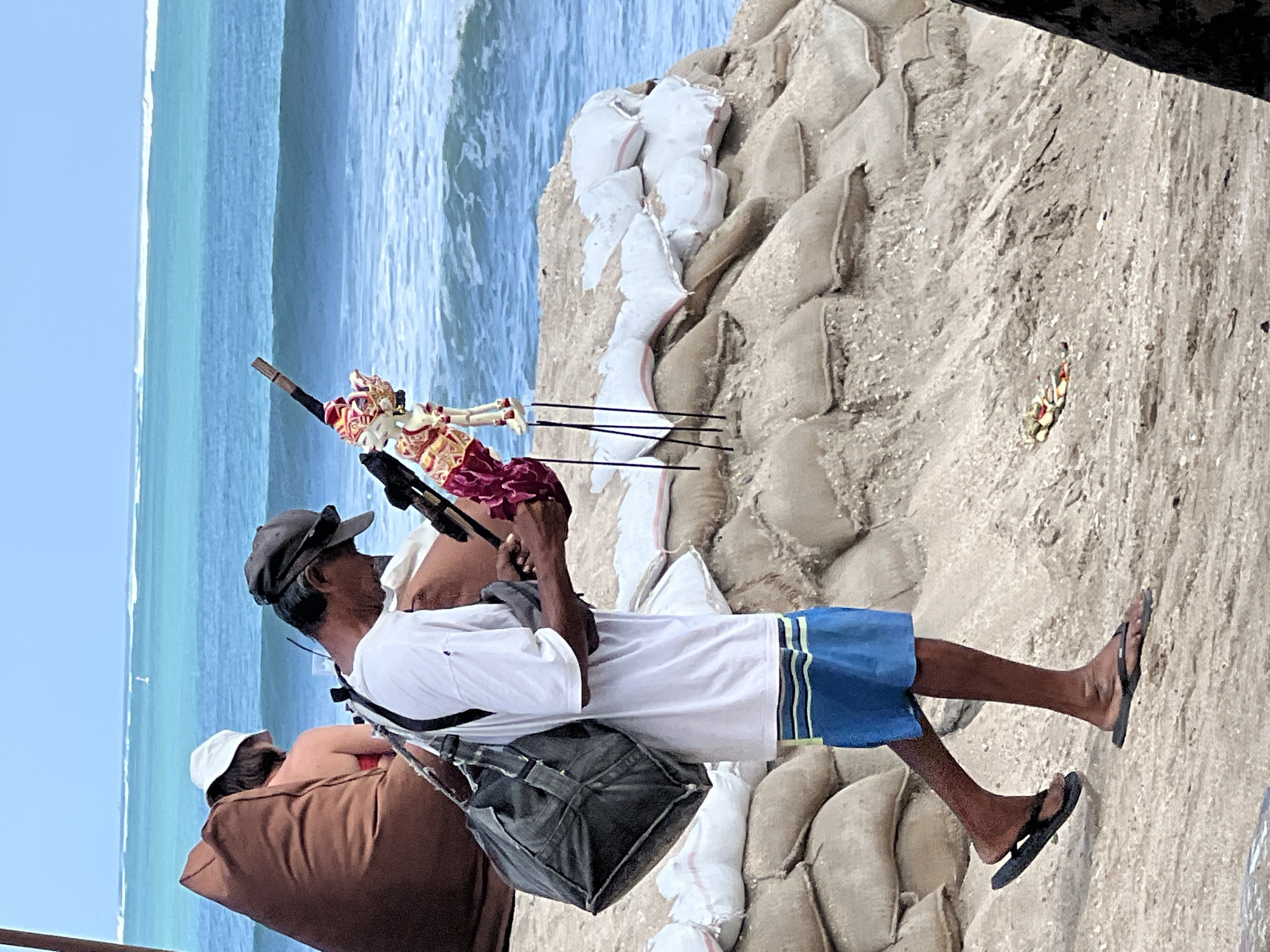 A man, appearing to be a street vendor, walks along a sandy beach in Kuta, Indonesia, holding a traditional Balinese Wayang puppet. The man is wearing a white t-shirt, blue and yellow striped shorts, and black flip-flops. He has a dark bag slung over his shoulder and a dark baseball cap on his head. The puppet, which is adorned in red and gold attire, is attached to a wooden stick. In the background, the turquoise ocean meets the horizon under a clear blue sky, with waves rolling in. The beach is lined with sandbags, likely for coastal protection, and a large brown beanbag chair is visible, where a person in a white hat and red bikini is seated. The overall scene depicts a moment on a popular beach, characterized by local culture and coastal scenery.