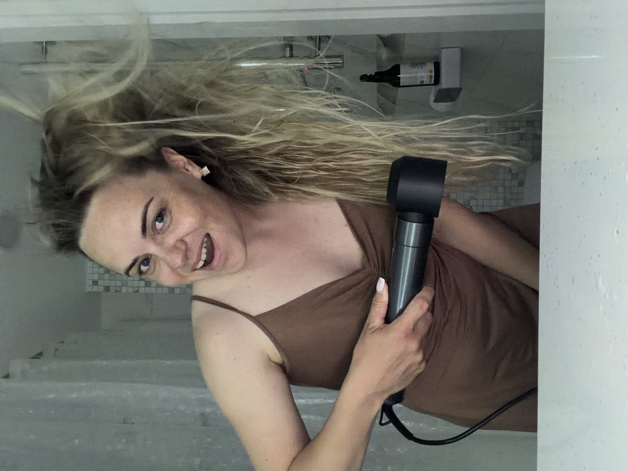 A woman with blonde hair, possibly in the middle of drying it, is holding a black hair dryer in her right hand. She is wearing a brown tank top and is smiling at the camera. The background appears to be a bathroom with gray-toned walls and a shower area. A shelf with a bottle and a box is visible on the wall. The location is uncertain but could possibly be Votkinsk, Russia.