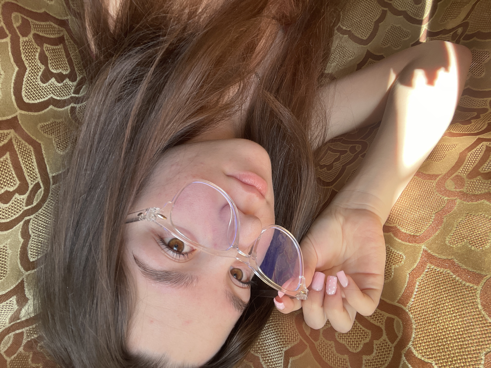 A young woman with long, brown hair is lying down on a textured, patterned surface, possibly a bedspread or cushion. She is wearing clear-framed glasses and gazing towards the camera. Her right hand, with pink nails, is holding the glasses slightly away from her face. Part of her arm is illuminated by a bright light source. The setting appears to be indoors. There is no evidence to indicate the location of Ольховка, Russia.