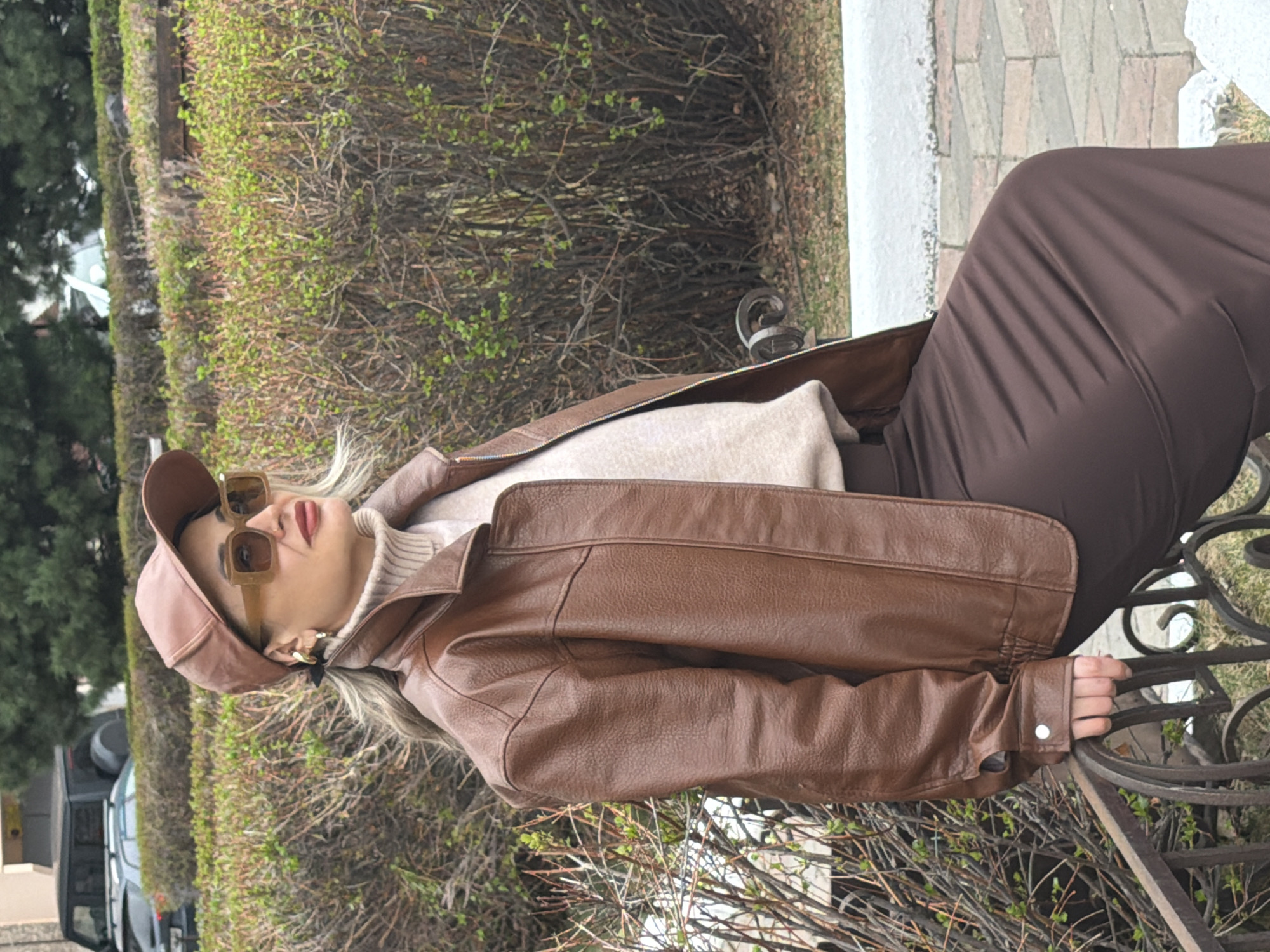 A woman is sitting on a wrought-iron bench in an outdoor setting. She is wearing a brown leather cap, brown-tinted sunglasses, a light beige turtleneck sweater, a brown leather jacket, and a long brown skirt. Her hand is resting on the bench's ornate metalwork. In the background, there are trees, shrubs with budding green leaves, and a paved path. A car is visible in the background. The exact location within Krasnoyarsk, Russia, cannot be determined from the image alone.