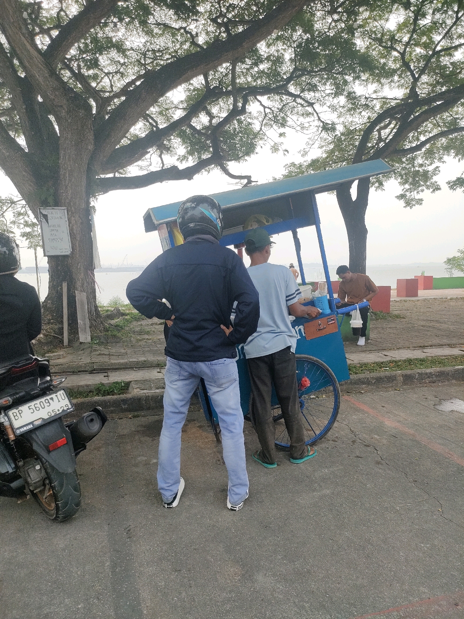 A low-angle, full shot captures a street scene at the waterfront in Batam, Indonesia. In the foreground, a motorcycle is parked on an asphalt road. The motorcycle's license plate reads "BP 5609 IU 05-29". To the right of the motorcycle, a person wearing a dark jacket and light blue jeans stands with hands on hips, facing a blue food cart. Behind the food cart, another person in a light blue t-shirt and dark pants stands. A third person in a brown shirt and black pants is seen in the background, holding a white plastic bag. The food cart is under a large tree with a sign attached to its trunk. In the distance, a body of water is visible, with a hazy sky and cranes on the horizon. Red concrete blocks are arranged along the waterfront.