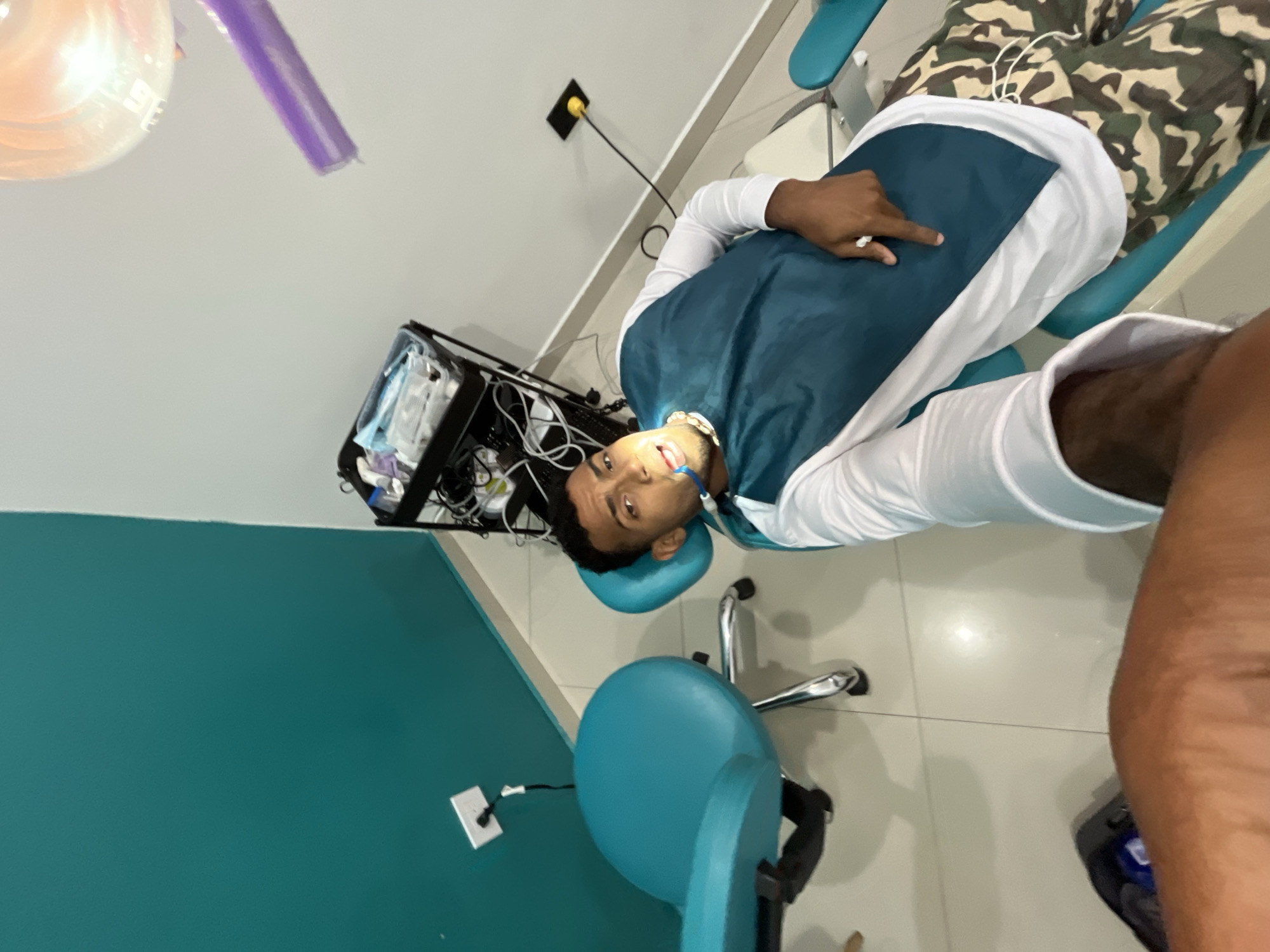 The image shows a man lying in a dental chair, presumably undergoing a dental procedure. He is wearing a teal dental bib over a white long-sleeve shirt and camouflage pants. A bright dental light is positioned above his face. Dental equipment and supplies are visible on a cart next to the chair. The room has a turquoise wall and light-colored flooring. A power outlet is visible on both the white and turquoise walls. The image does not provide sufficient detail to determine the exact location within La Asuncion, Venezuela.