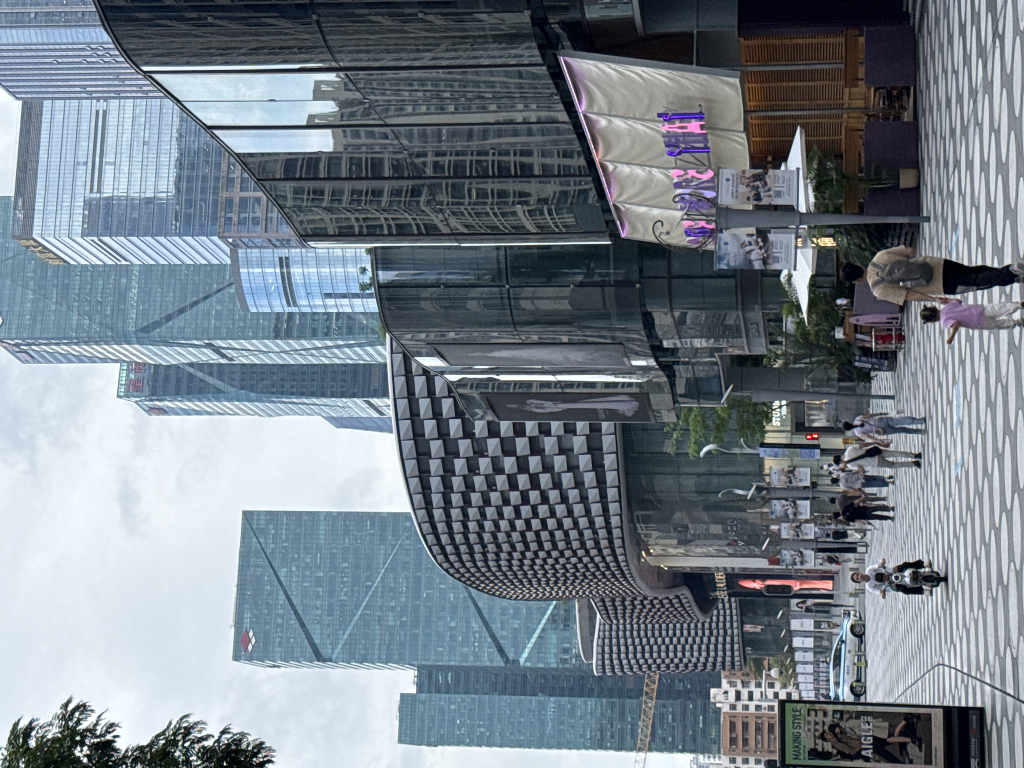 The image depicts a street scene in Futian District, China, characterized by modern architecture and pedestrian activity. Several skyscrapers with glass facades dominate the background. A building with a distinctive checkered pattern is also prominent. The street is paved with a patterned design and features a line of streetlights. Several pedestrians are walking along the street. Signage for various businesses is visible, including a sign that says "MAKING STYLE AIGLE." A blue car is parked. The sky is overcast.