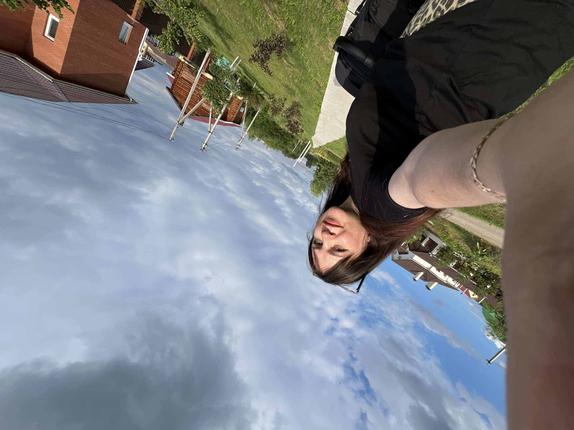 A woman is taking a selfie outdoors in Noviy, Russia. She is wearing a black t-shirt and a gold bracelet. Her arm is in the foreground, extended towards the camera. In the background, there are several houses with red and brown roofs, green grass, and trees. The sky is partly cloudy with a mix of dark and light clouds. There are also power lines visible in the background. The image is taken at an angle, with the woman's head tilted slightly.