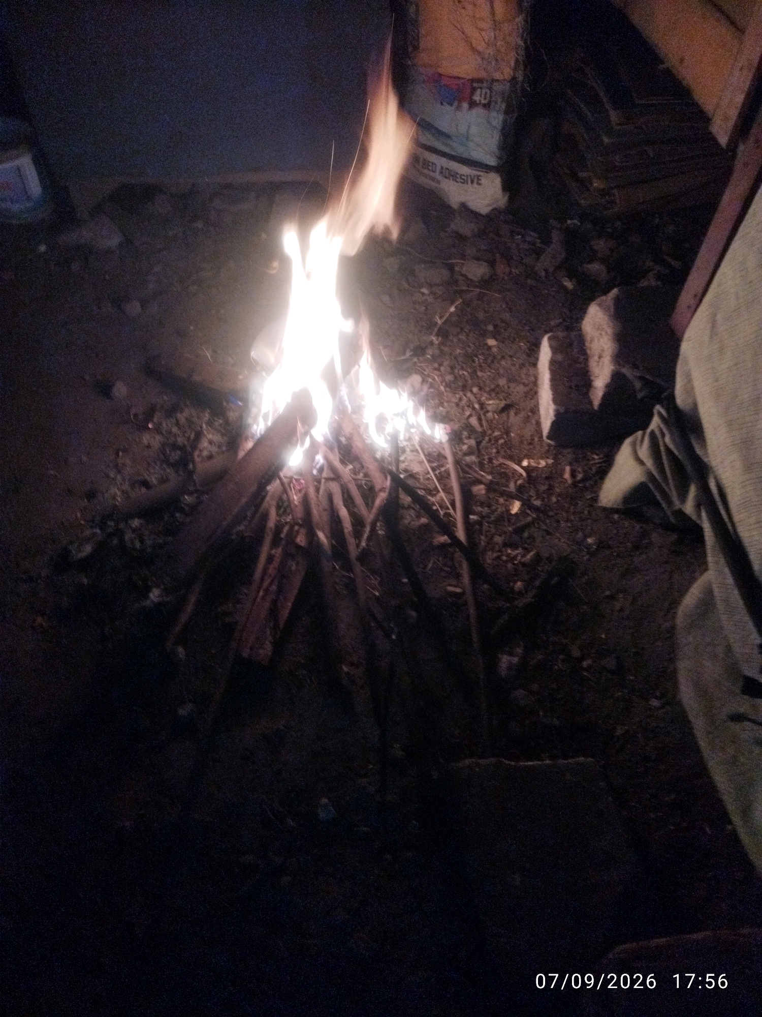 A campfire burns brightly at night in an outdoor setting. The fire is made of stacked logs and branches, with flames reaching upwards. The ground around the fire appears to be dirt and rubble, with scattered rocks. In the background, there is a partially visible bag with "BED ADHESIVE" and the number "40" printed on it. To the right of the fire, a person's leg and part of their clothing, a light-colored fabric, are visible, suggesting they are sitting or standing near the fire. The timestamp "07/09/2026 17:56" is overlaid on the bottom right corner of the image. The overall scene is dimly lit, with the fire being the primary light source. The location appears to be an unpaved, possibly industrial or construction area.