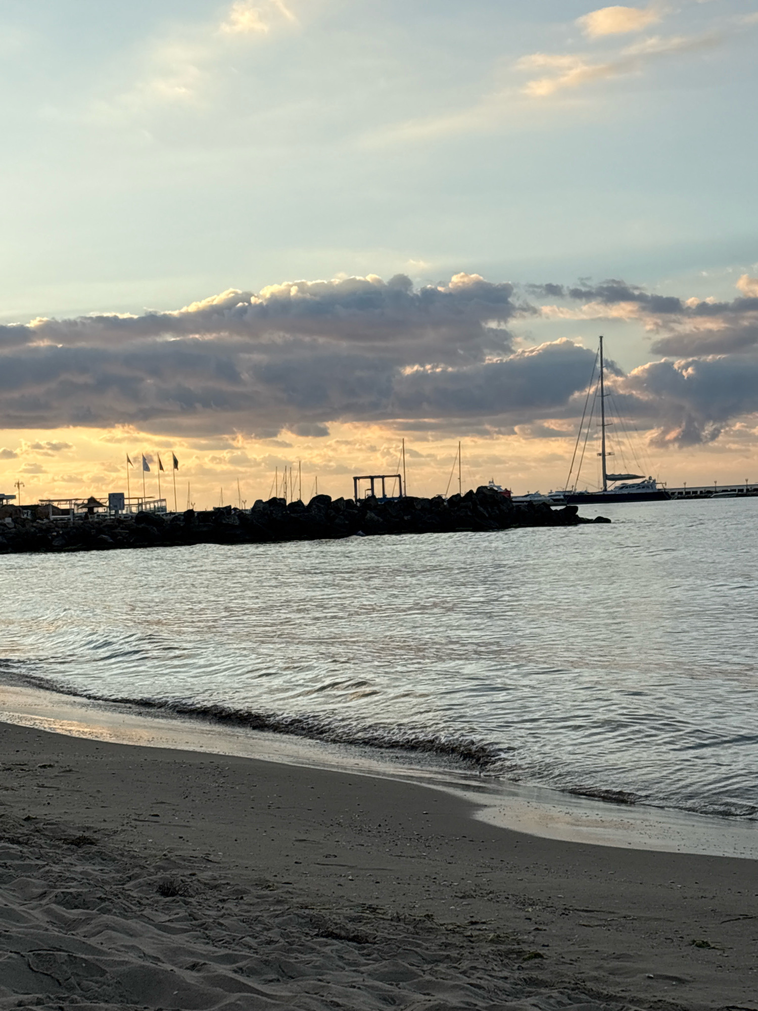 A beach scene at sunset is depicted, featuring a calm sea with gentle waves lapping onto the shore. In the foreground, dark sand is visible, marked by tire tracks. The middle ground showcases a breakwater made of dark rocks, with several flagpoles flying flags, structures, and moored sailboats. A prominent sailboat with a tall mast is anchored further out in the water, silhouetted against the vibrant sky. The sky itself is filled with dramatic clouds, illuminated by the warm hues of the setting sun. The scene is located in Sveti Vlas, Bulgaria.