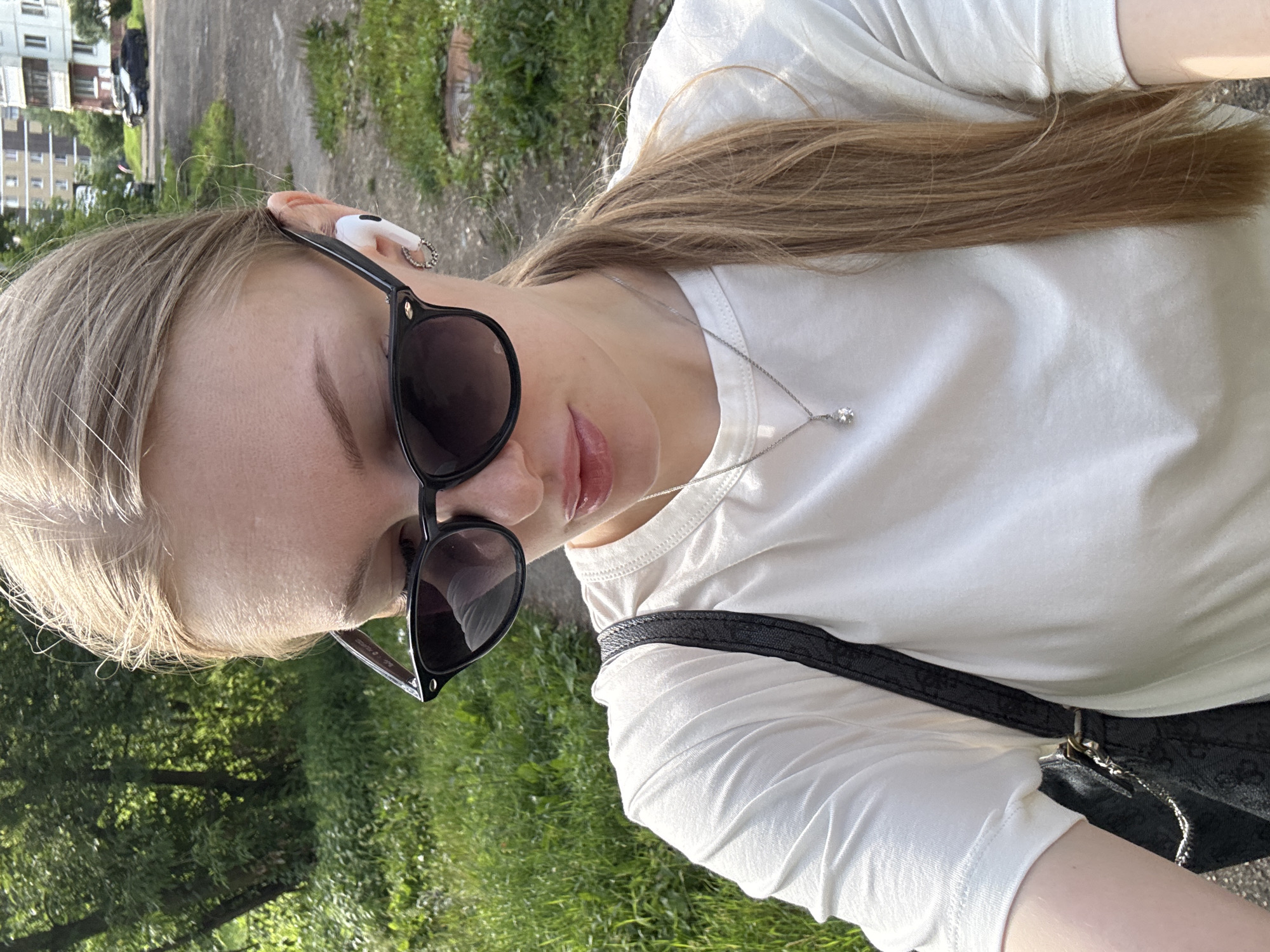 A young Caucasian woman with blonde hair pulled back in a ponytail and wearing sunglasses, a white t-shirt, and a silver necklace is seen from the chest up, outdoors in the city of Perm, Russia. She has a white wireless earbud in her right ear and is wearing a small hoop earring. The woman is looking down and to her left, with her mouth slightly open. She is carrying a black shoulder bag with a chain strap. The background shows a grassy area with trees and a building with multiple windows and balconies, likely an apartment building. Two cars are visible parked near the building, and a manhole cover is partially visible in the grass to the right. The lighting suggests it is daytime, likely late morning or afternoon, with sunlight casting shadows.