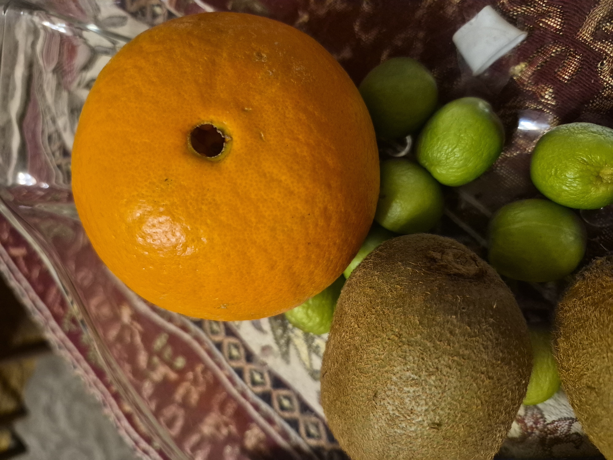 The image displays an overhead close-up view of various fruits arranged in a clear container, resting on patterned textiles. A prominent, vibrant orange citrus fruit is positioned on the left, featuring a distinct, circular hole on its top surface. To the right and behind the orange are several small, bright green limes. Below the orange and limes, at least two fuzzy, brown kiwi fruits are visible, partially obscuring each other. The fruits are held within a transparent material, likely plastic or glass, which exhibits reflections and distortions. Beneath the container, two different patterned fabrics are visible: one with geometric diamond shapes in shades of brown and white, and another primarily maroon and red with golden or light-colored intricate designs. A small, crumpled white object, possibly paper, is seen among the limes in the upper right. There is no discernible activity taking place, nor any visible text, specific time of day, weather, or human emotions. The scene appears to be an indoor setting. The location context provided is Yerevan, Armenia.