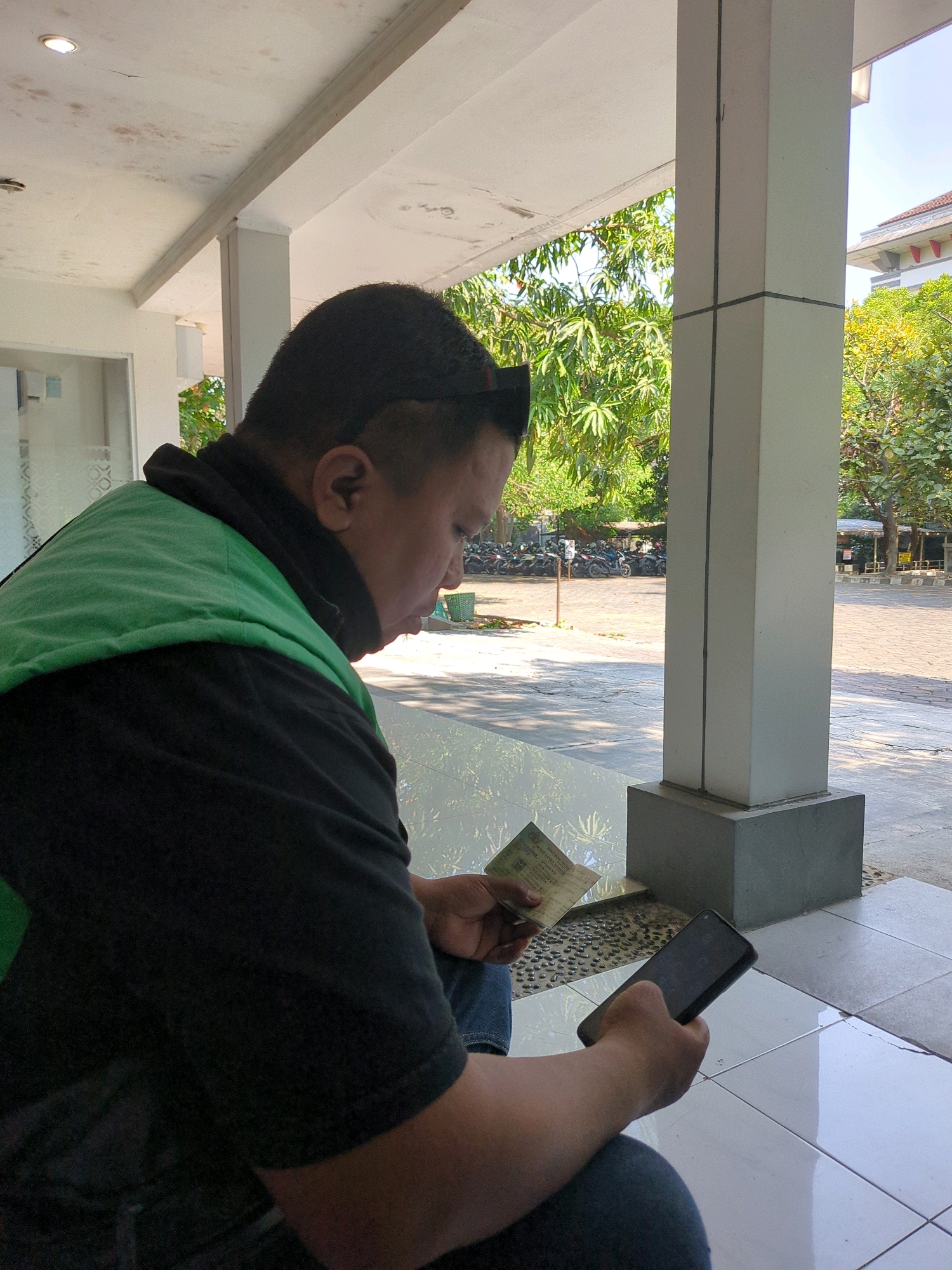 The image shows a man sitting outdoors in Surakarta, Indonesia, looking down at a document and a smartphone. He is wearing a black shirt and a bright green vest. The man is positioned in the foreground, with his right shoulder and side facing the camera. He holds a small green booklet in his left hand, and a black smartphone in his right hand.
The background reveals a paved area with several motorcycles parked. Trees and foliage are visible behind the parking lot, and a building with a reddish roof can be seen in the distance. The scene appears to be an outdoor waiting area or public space, possibly near a facility or establishment in Surakarta. The lighting suggests it is daytime, with sunlight casting shadows. The man's posture and focus on the items in his hands indicate he is engaged in reading or interacting with his phone.