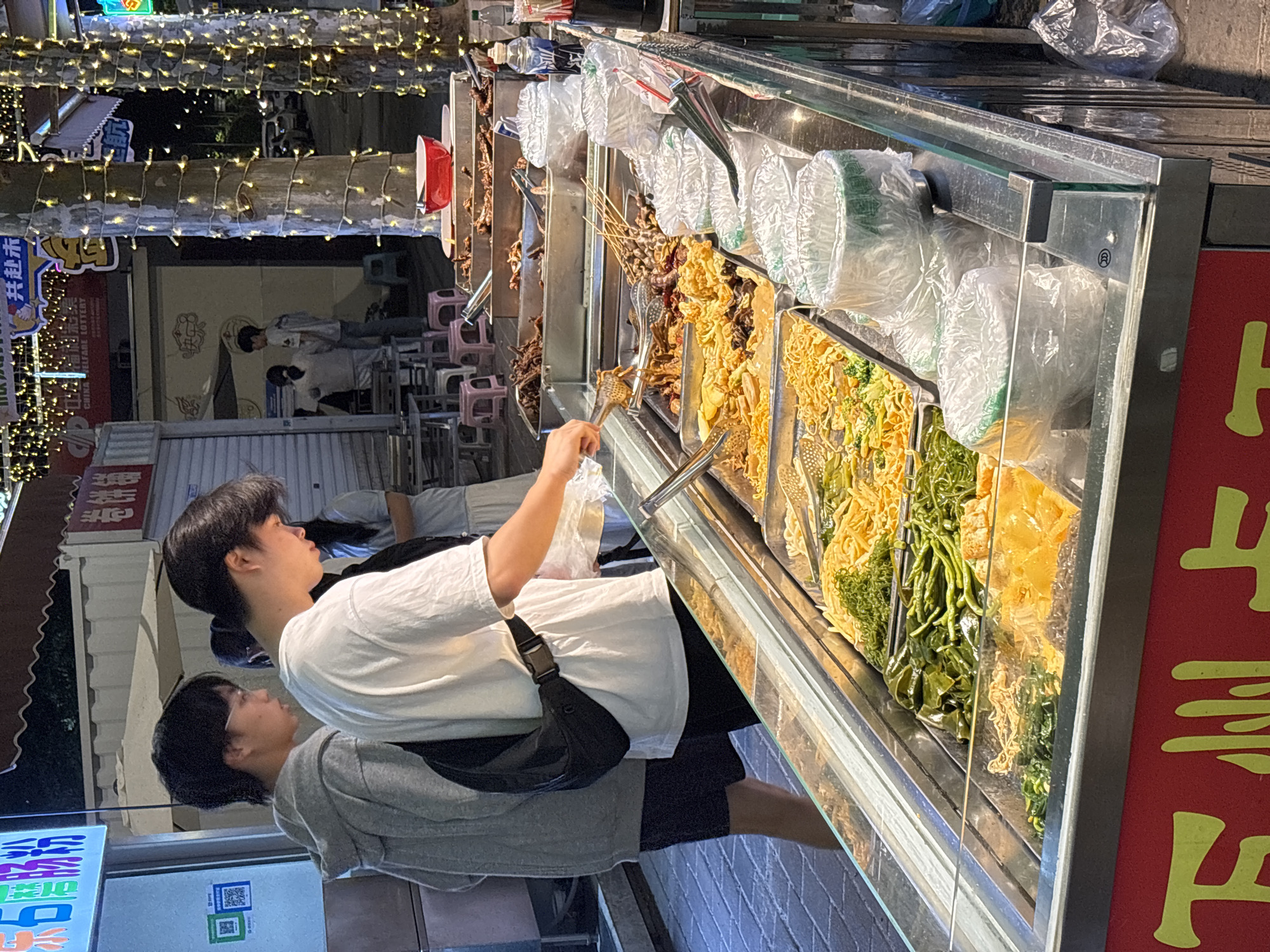Here is a description of the image:
A street food vendor is seen at night in the Jiangjin district of Chongqing, China. The view is from a slight angle. A young man wearing a white t-shirt and a black shoulder bag is using tongs to select food from a display case. The food display case is filled with skewers of various cooked foods. The vendor stall is well-lit with many strung lights overhead. Other patrons are standing behind the young man. There are Chinese characters on the signage of the food stall.