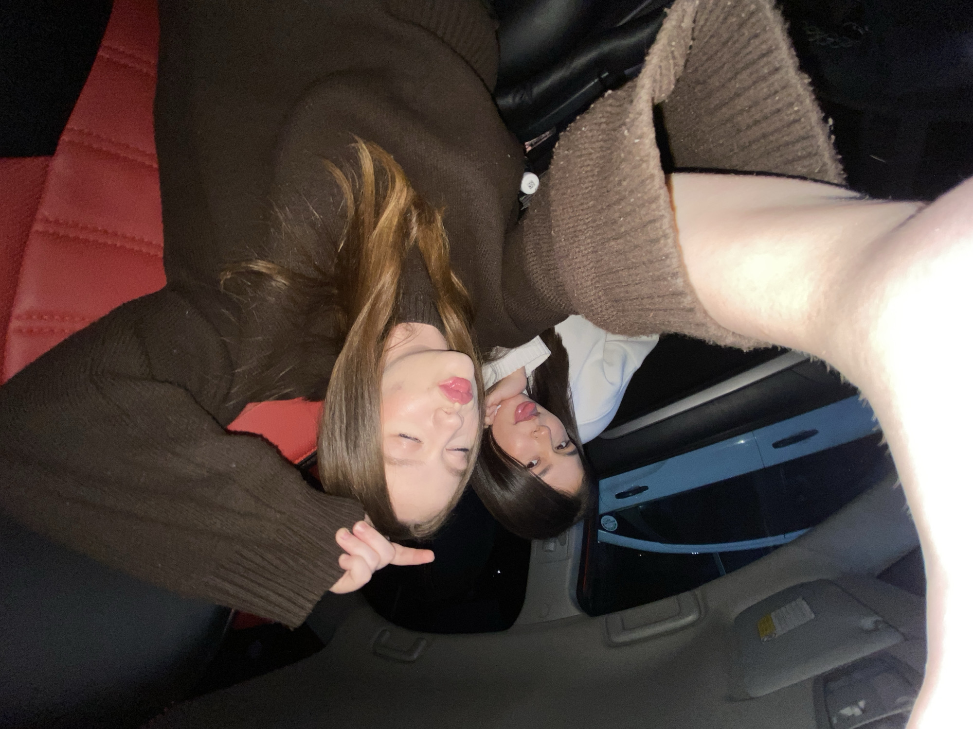 Two young women are taking a selfie inside a car, possibly in Chaikovsky, Russia, though there is no explicit confirmation of the location in the image. The woman on the left has light brown hair and is wearing a dark brown sweater, pointing two fingers. The other woman has dark hair and is wearing a white top. Red leather car seats are visible. The car has a sunroof.