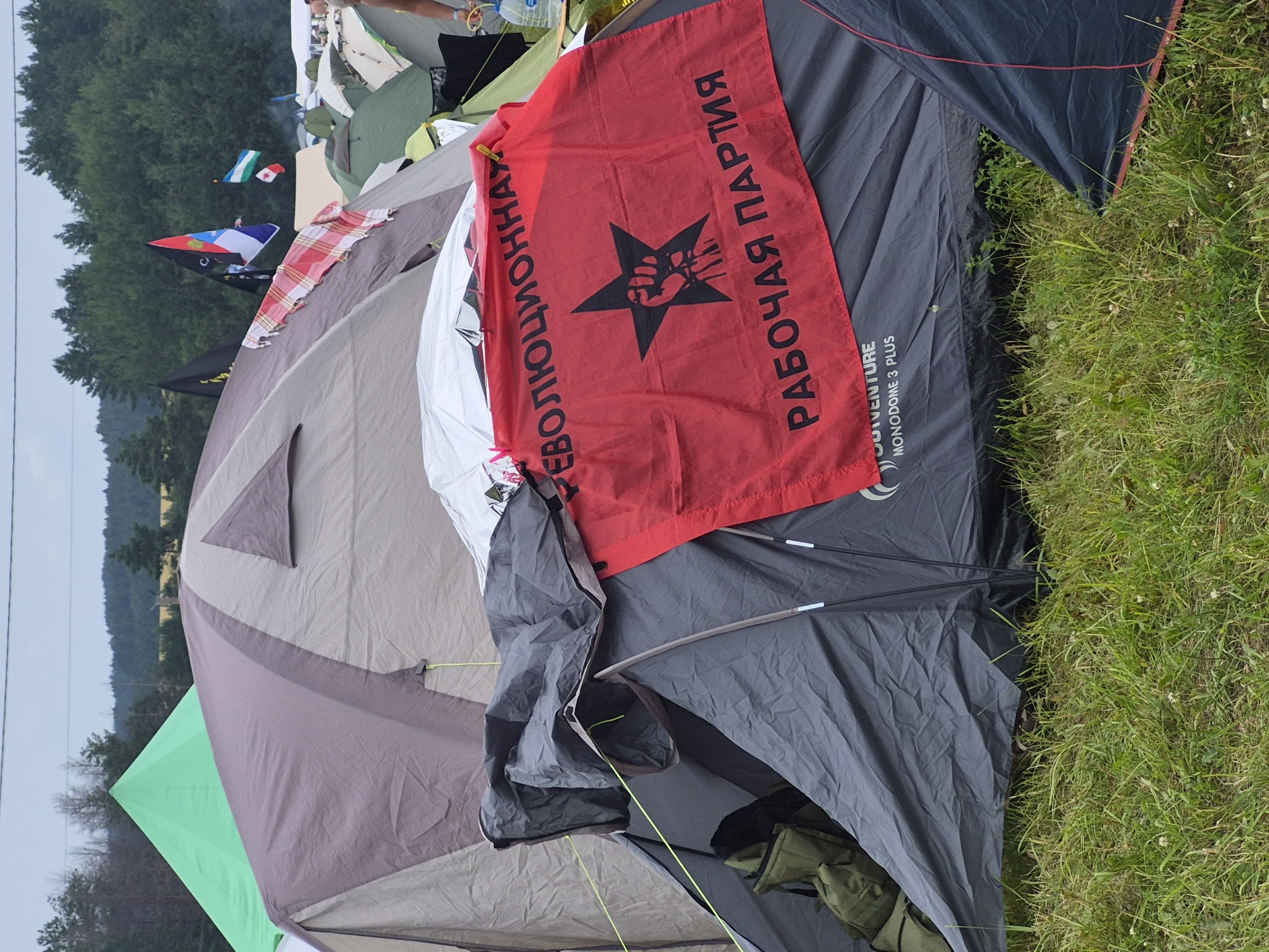 The image shows a campsite with several tents. A prominent red flag with a black star and a fist symbol is draped over a gray tent. The flag has Russian text that reads "РЕВОЛЮЦИОННАЯ РАБОЧАЯ ПАРТИЯ" (Revolutionary Workers' Party). In the background, there are trees and other tents, as well as several flags. The ground is covered in grass. The scene appears to be an outdoor event or festival. There are no people clearly visible in the foreground. The location is identified as Lagunovo, Russia.