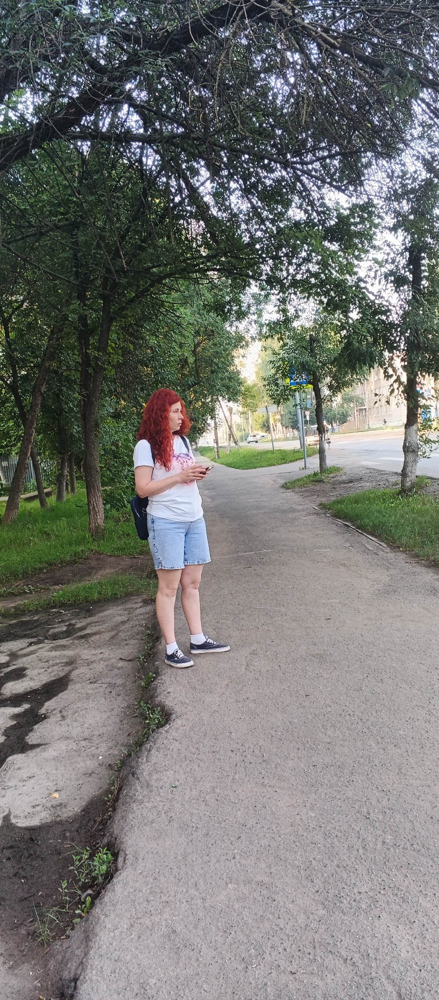 A Caucasian woman with long, curly red hair stands on a sidewalk in Izhevsk, Russia. She is wearing a white t-shirt, light blue denim shorts, and dark sneakers with white socks. She is holding a mobile phone in her hands and looking to her right. A dark backpack is slung over her left shoulder. The sidewalk is paved with concrete, and to the left of the sidewalk, there is a grassy area with trees. To the right of the sidewalk, there is a street with some buildings visible in the background. The lighting suggests it is daytime, possibly late afternoon or early evening. There are no other people or animals in the immediate vicinity.