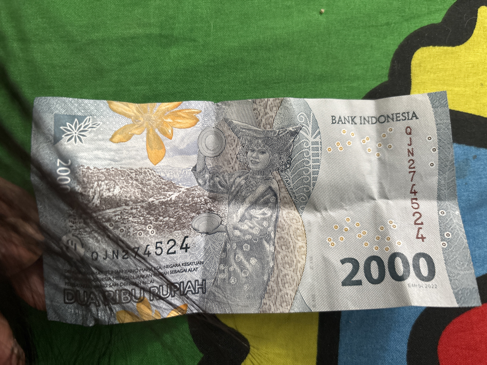 The image shows a 2000 Indonesian Rupiah banknote. The bill features an illustration of a woman in traditional clothing and headwear, along with the denomination "2000", the words "BANK INDONESIA", and the serial number "QJN274524". The words "DUA RIBU RUPIAH" are also visible. The banknote is lying on a multicolored fabric with green, yellow, black and blue patterns. A dark object, resembling human hair, partially obscures the lower left area of the banknote. The bill is creased in the middle. There is no information in the image to associate it with North Jakarta.