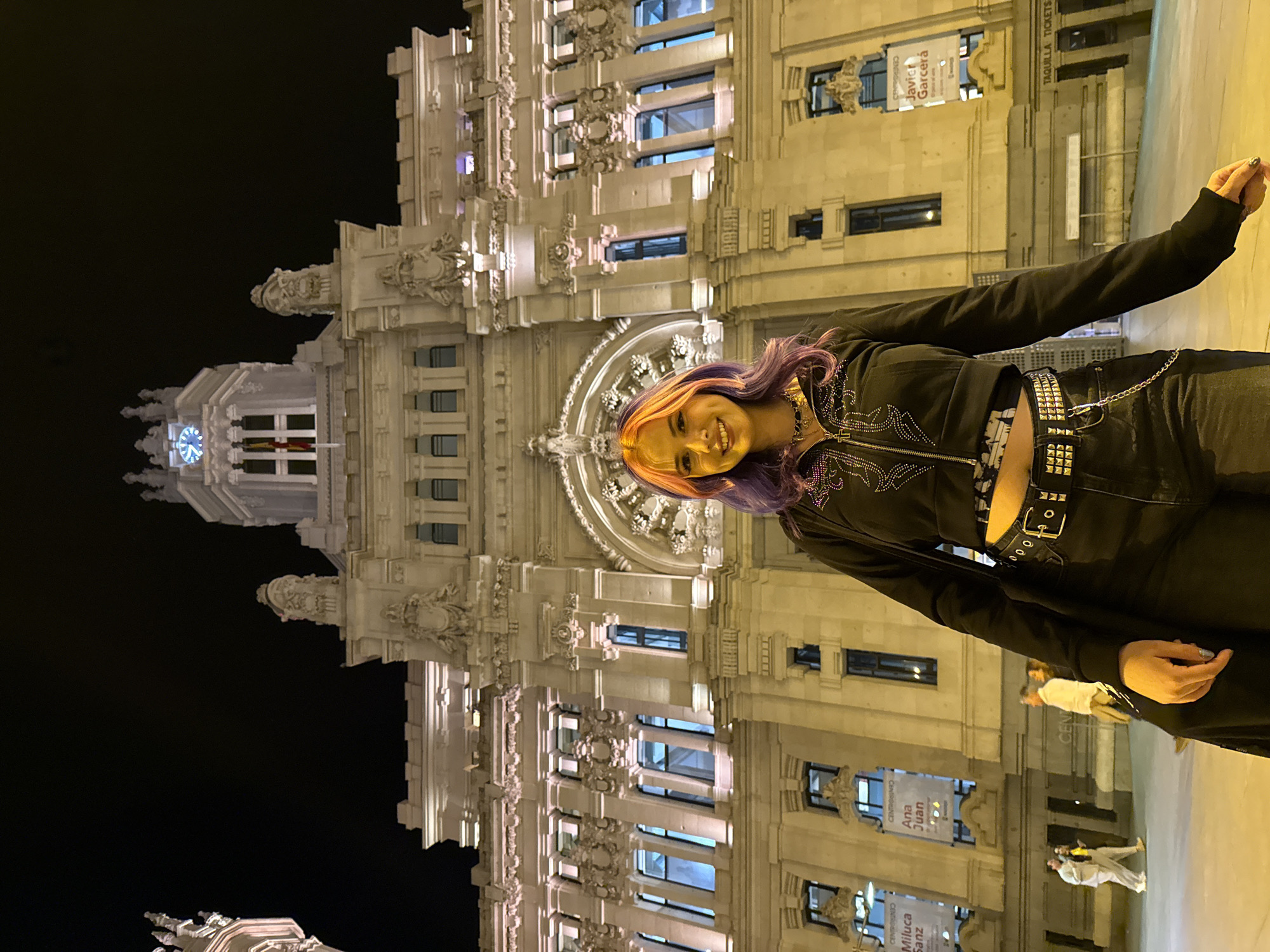 A nighttime, medium shot depicts a smiling individual with purple hair, a studded belt, and a black jacket standing in front of the Cibeles Palace in Madrid, Spain. The Palace is illuminated with the night sky in the background and various people in the foreground.