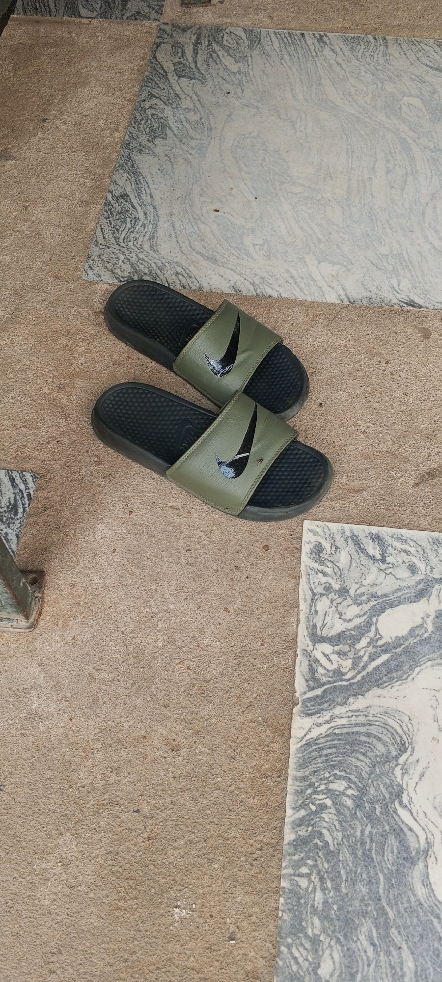 A pair of olive green Nike slide sandals are positioned on a sandy-colored surface, possibly concrete or dirt, with two large, patterned stone slabs visible to the left and right. The sandals are dark green with a black Nike swoosh logo on each strap and a textured black sole. They are lying flat, side-by-side. The stone slabs have a marbled appearance with swirling patterns of gray and white. The overall setting appears to be outdoors, on a paved or ground-level surface, possibly in Jalingo, Nigeria. There are no people present in the image.