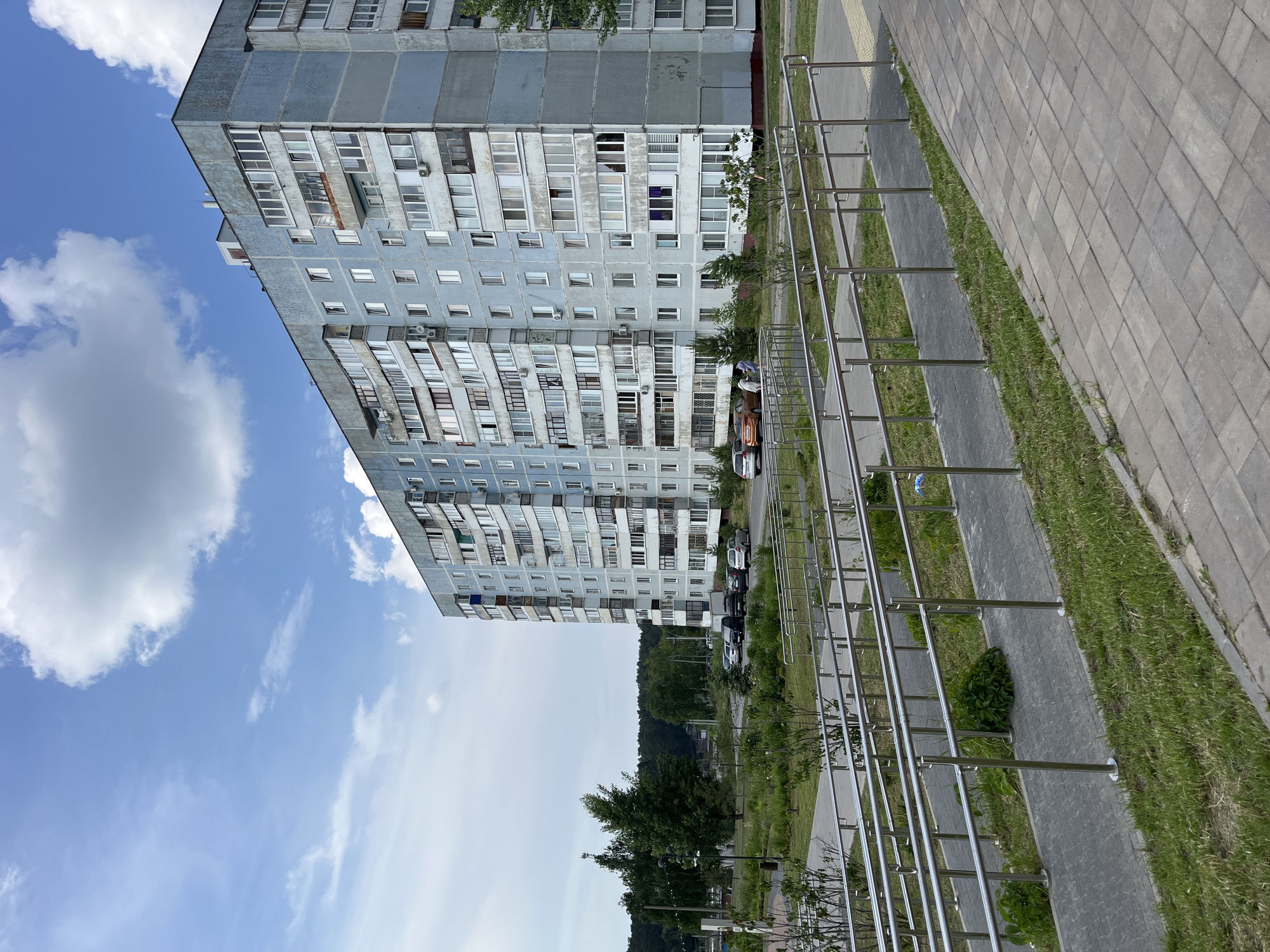 An outdoor, eye-level, medium shot captures a light blue, multi-story apartment building in Zelenodol'sk, Russia, under a cloudy blue sky. The building, a typical Soviet-era residential structure, exhibits signs of age with some exterior wear. In front of the building, a newly paved walkway with metal handrails guides pedestrians. A patch of green grass and small bushes borders the walkway. Beyond the building, a dense forest provides a natural backdrop. Several cars are parked in front of the building, and two individuals are seen walking near them. The scene suggests a residential urban environment on a pleasant day.