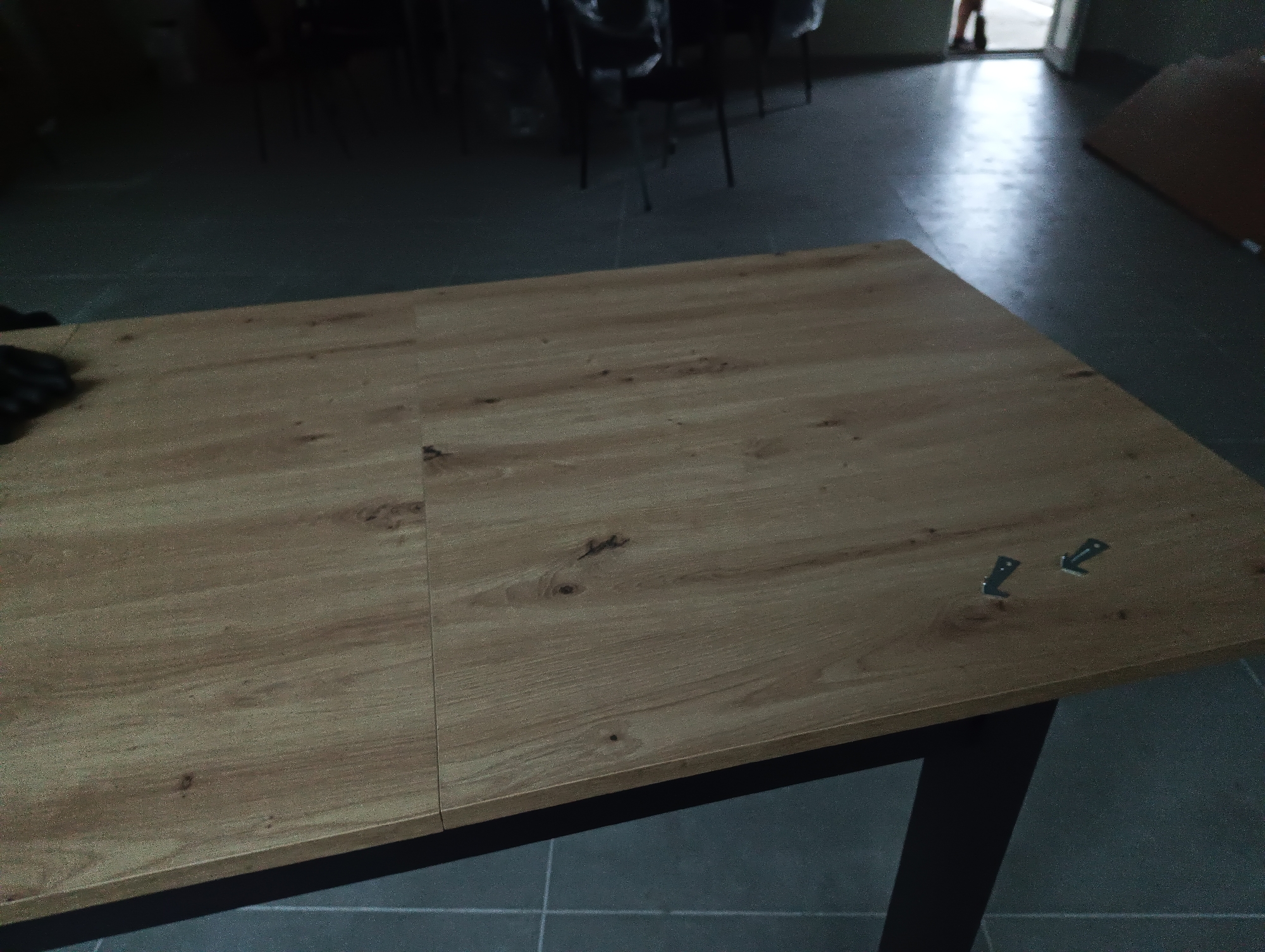 The image captures an indoor scene, dominated by a large, rectangular table with a light-brown wooden top. The table surface features a distinct seam running down its length, suggesting it might be an extendable table or constructed from two main sections. The wood grain is visible, with occasional darker knots.
On the far left edge of the table, a dark-gloved hand is partially visible, resting on the surface. Towards the right side of the table, two small, metallic, L-shaped brackets or clips are loosely placed. A dark, possibly metal, table leg is visible in the bottom right corner of the frame.
The background is dimly lit and somewhat blurry. To the left and center-back, dark, indistinct shapes appear to be chairs or other furniture, some with thin legs. To the right, in the background, a bright doorway illuminates a section of a dark, tiled or concrete floor. A person's lower legs and feet are visible standing in this doorway, suggesting they are either entering or exiting the room. Further right, a large, reddish-brown rectangular object, possibly a wood panel, leans against something on the floor.
The overall impression is that of a functional space, possibly a workshop or assembly area, where furniture is being set up or put together. The presence of the loose brackets and the gloved hand on the table reinforces this idea of assembly or preparation. There is no visible text in the image.