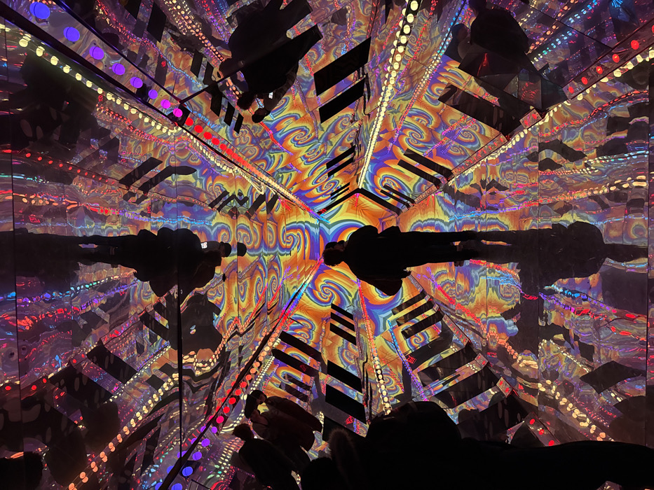 A vibrant, psychedelic tunnel of swirling orange, yellow, blue, and purple patterns is reflected in mirrored surfaces. Black geometric shapes break up the visual flow. Silhouettes of people are positioned at the bottom and top of the frame, their forms duplicated in the kaleidoscopic reflections. Strings of colored lights line the edges of the mirrored panels, enhancing the trippy, immersive effect of the space.