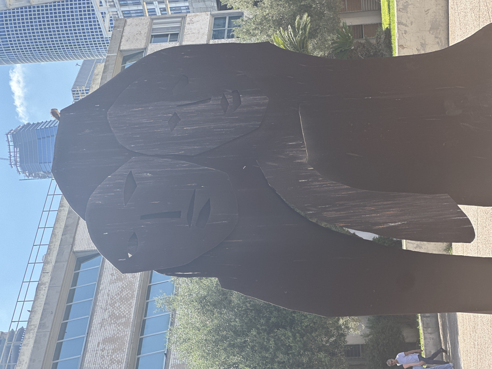The image shows a large, abstract metal sculpture with two stylized faces in the foreground. The sculpture is dark brown and appears to be made of weathered metal. In the background, there are modern buildings, some with glass facades, and trees. A bird is perched on top of the sculpture. To the left, two people are walking on a paved walkway in front of a low concrete wall and some greenery. The scene appears to be in a public space within Tel Aviv, Israel.