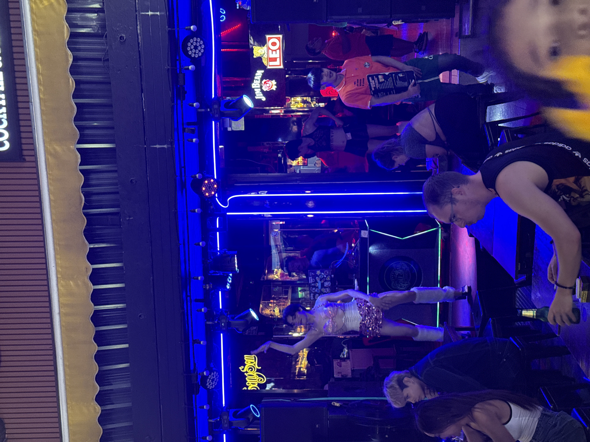 The image shows the interior of a bar in Bangkok, Thailand. The bar has bright blue neon lighting and features a stage. On the stage, a performer in a white and pink sequined outfit is dancing. Several patrons are in attendance, seated at tables. One patron is standing near a table, holding a bottle of beer. Beer brands like "Leo" and "Jim Beam" are visible in the background, suggesting alcohol is served at the establishment. A waiter is seen holding a menu. The awning and sign reading "Cocktail" indicate the establishment serves alcoholic beverages.