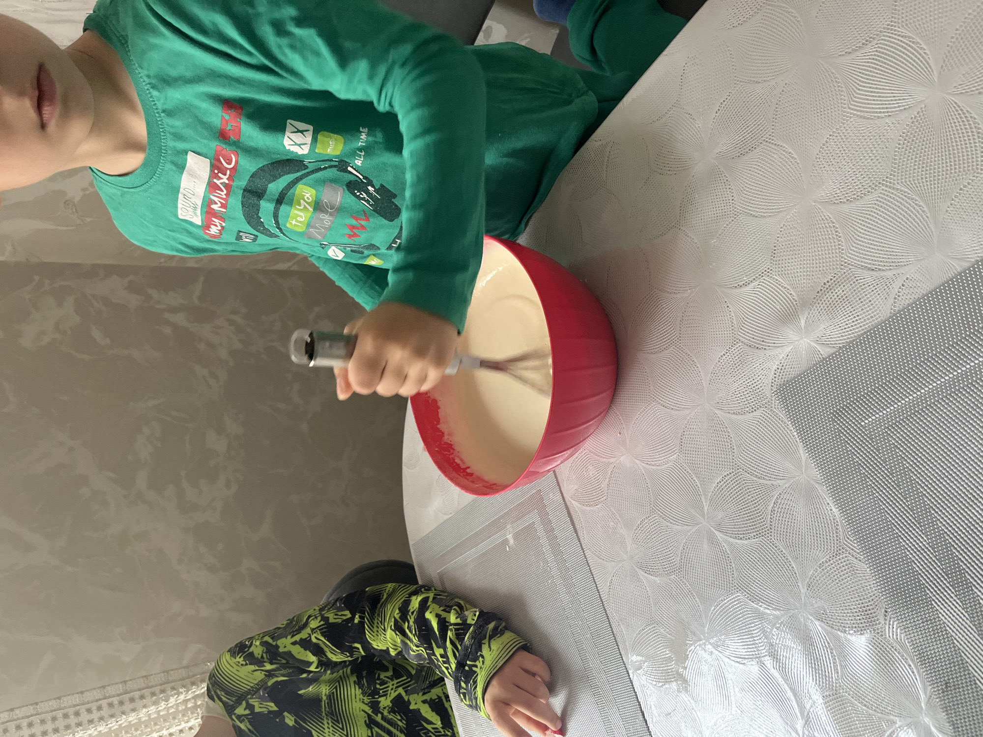 A child in a green long-sleeved shirt is whisking batter in a red bowl on a white tablecloth with a floral pattern. Another child, wearing a green and black patterned jacket, is partially visible at the bottom of the frame. The photo was taken in Kaspiysk, Russia, inside a home. The walls are a light marble pattern.