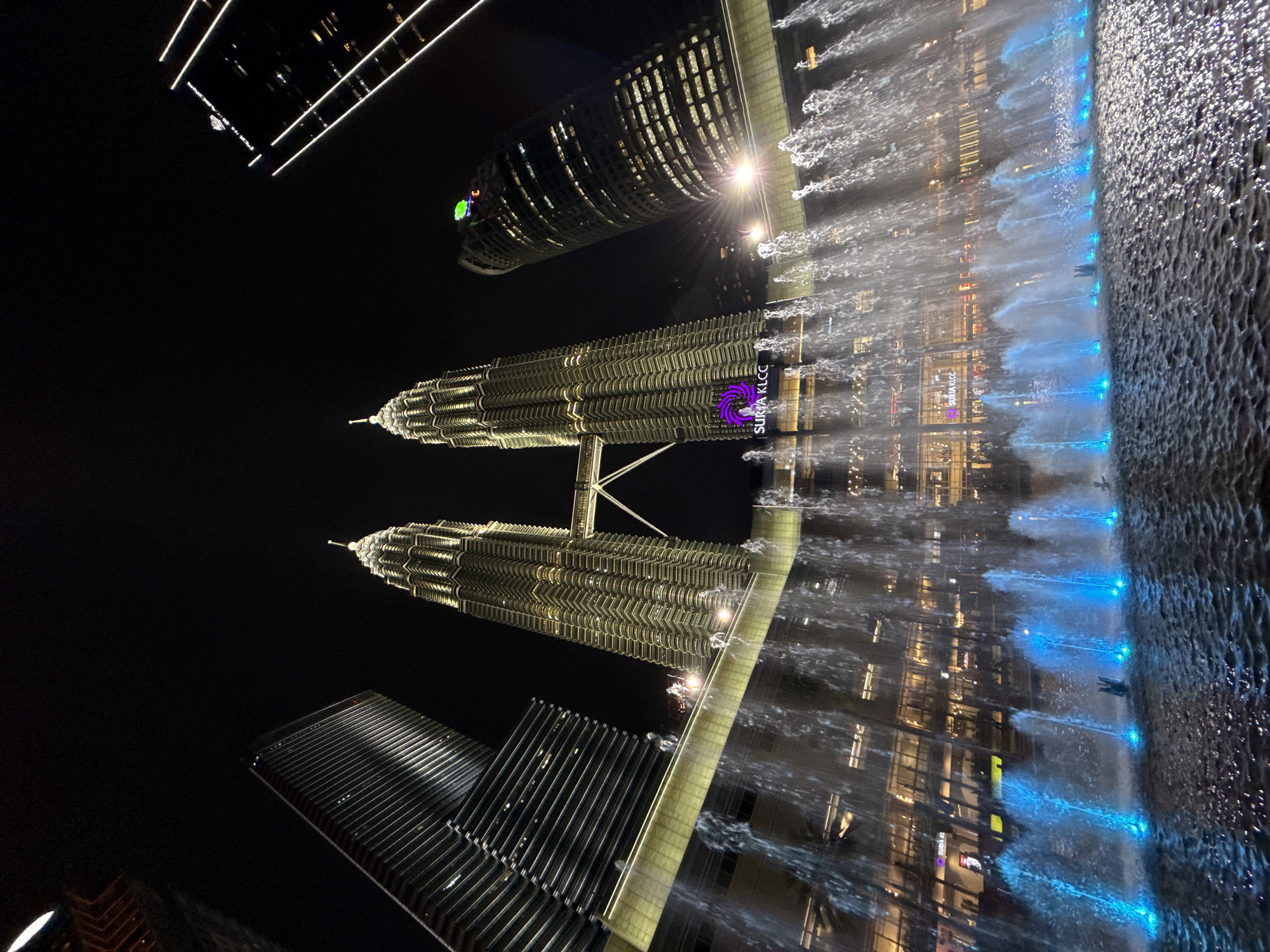 The image depicts a nighttime view of the Kuala Lumpur City Centre (KLCC) in Kuala Lumpur, Malaysia. Prominently featured are the Petronas Twin Towers, illuminated in yellow light, with the Suria KLCC shopping mall situated at their base. A water fountain is visible in the foreground with blue underwater lights, reflecting the surrounding buildings. Several other skyscrapers, including a cylindrical-shaped tower, are also present in the frame. The image captures a dynamic interplay of architectural and aquatic elements in an urban setting.