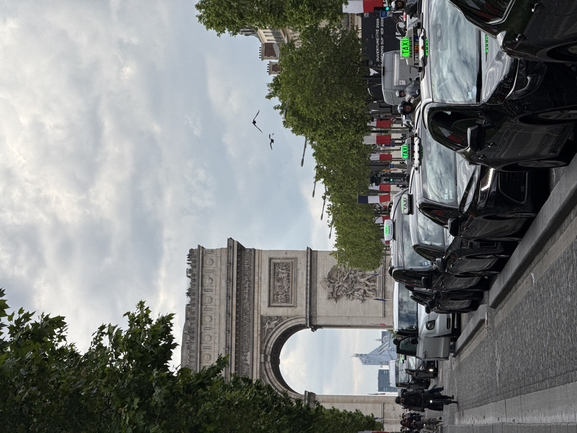 A street-level, slightly skewed photograph shows the Arc de Triomphe in Paris, France. A row of black taxis, identified by their "TAXI" signs, are parked on the right-hand side of the frame. Trees line the street, and the sky is overcast. Birds are visible in the sky above the Arc. People and motorcycles are at the front, near the curb.