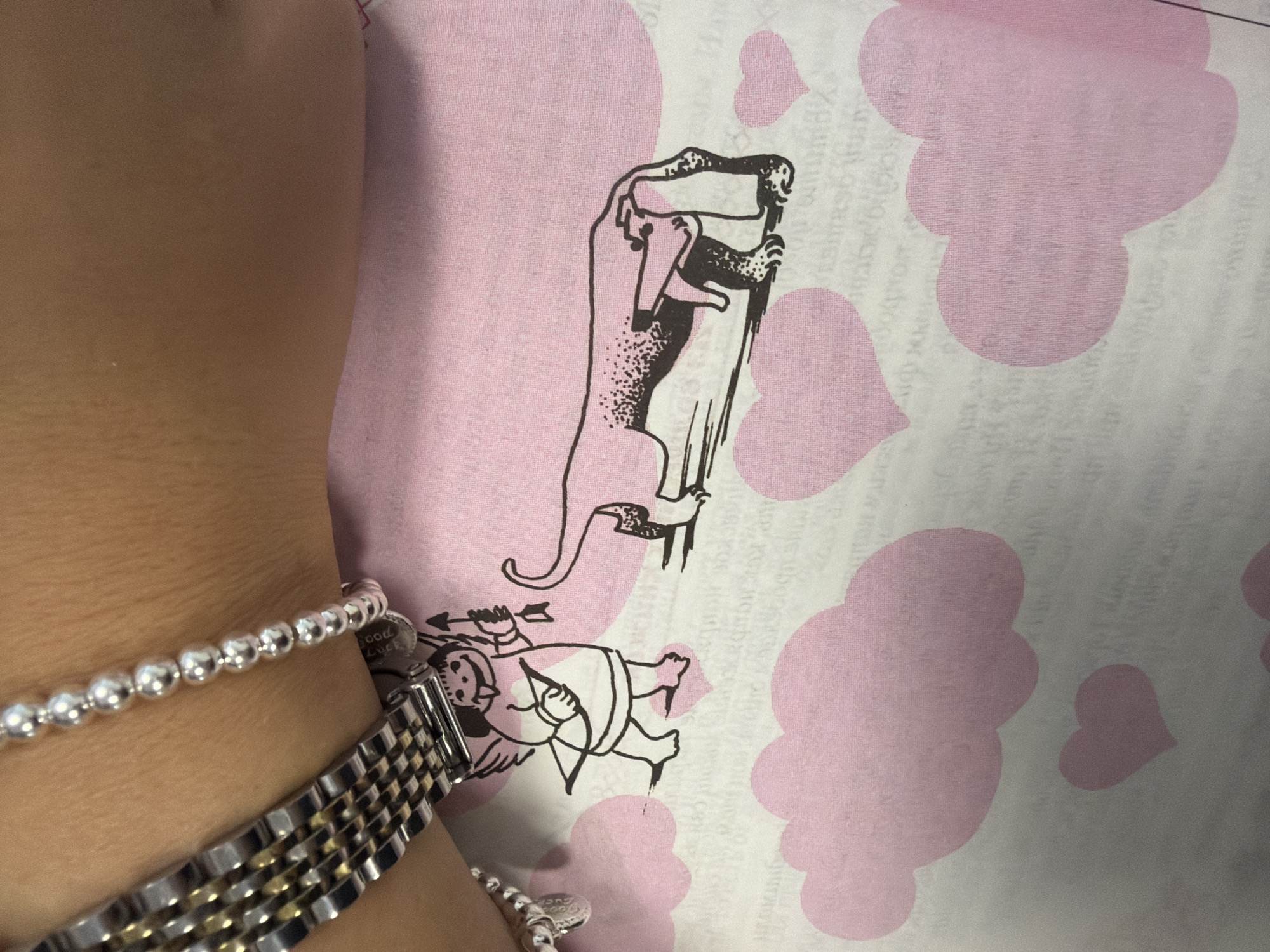 The image shows a close-up of a person's arm with two bracelets. One bracelet is made of small silver beads, and the other appears to be a silver metal watch band with gold accents. The arm is resting on a pink and white patterned fabric. The pattern includes stylized clouds and hearts. There is also a black and white illustration of a cherub holding a bow and arrow, and a long, thin dog-like creature. The text on the fabric is partially visible and appears to be in a foreign language, potentially Cyrillic script. There are no recognizable landmarks or contextual clues to indicate a specific location in Homyel, Belarus. The scene is an indoor setting, likely a desk or table, given the fabric is spread out.