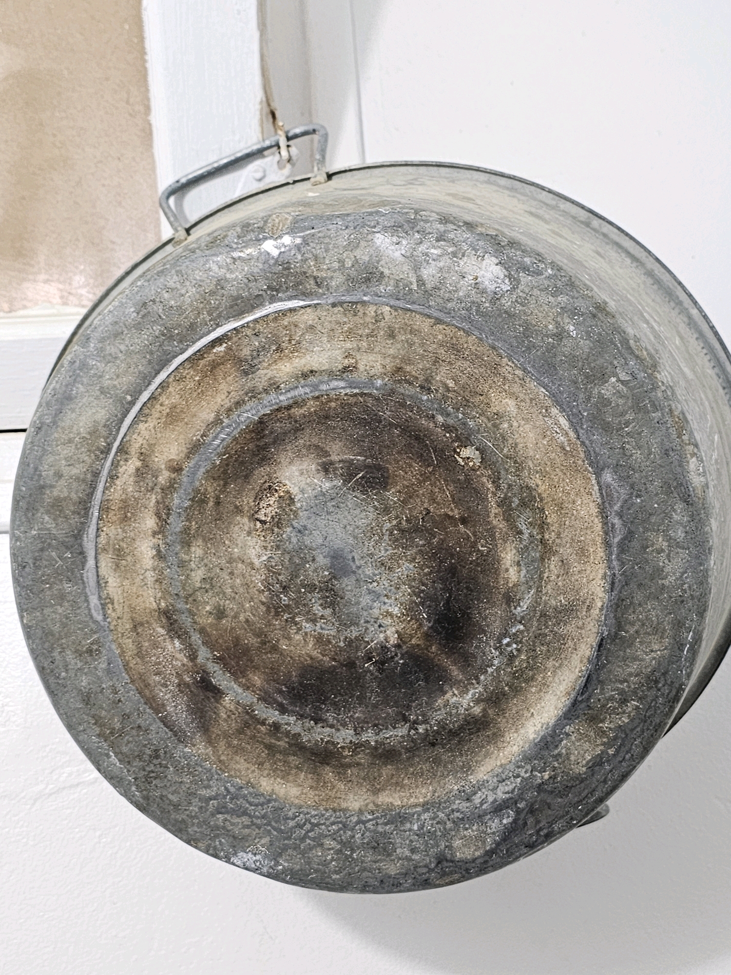 The image is a close-up, eye-level shot of the bottom of a galvanized metal bucket. The bucket appears old and weathered, with a textured, mottled surface. There are circular concentric rings on the base, typical of bucket construction, with darker, burnt-looking discoloration in the center. A metal handle is visible attached to the rim of the bucket at the top left of the frame. The bucket is hanging against a white wall, with a portion of a cream-colored window frame visible in the upper left background. The lighting is diffused, casting soft shadows. There are no people or animals present in the image. The scene is domestic or utilitarian, likely within a building. The image does not contain any information to indicate a specific location in Batna, Algeria.