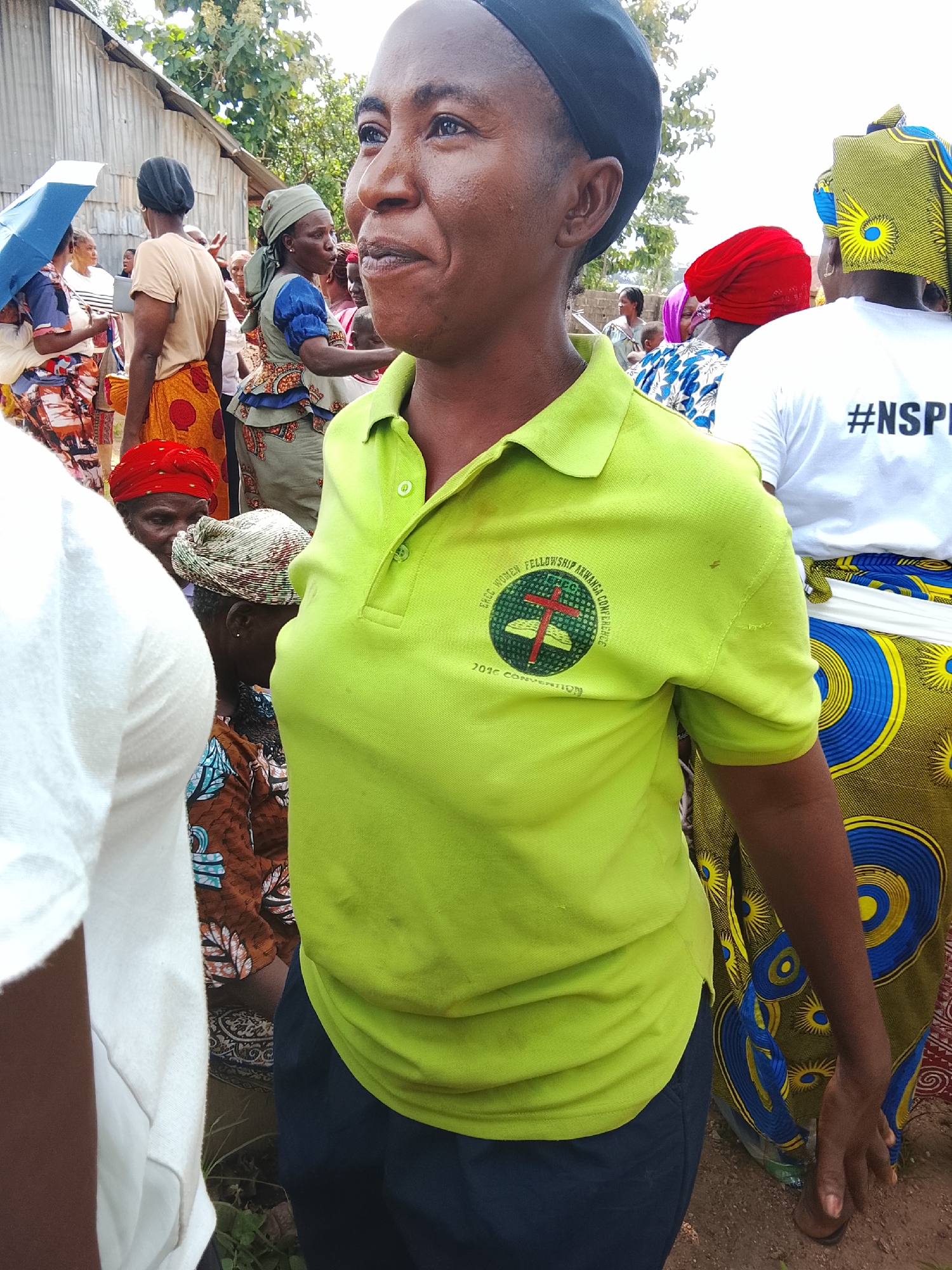 The image depicts an outdoor scene in Akwanga, Nigeria, where a group of people, primarily women, are gathered. In the foreground, a woman wearing a lime green polo shirt and dark pants is the central figure, looking to her right with a slight smile. The polo shirt has a circular emblem on the chest with text that appears to read "FREE WOMEN FELLOWSHIP AKWANGA CONFERENCE 2016 CONVENTION". Behind her, to the right, another woman is visible from the chest down, wearing a white t-shirt with the hashtag "#NSPI" printed on it, and a colorful patterned skirt. To the left and further back, several other individuals are present, some holding umbrellas and wearing various traditional African attire and headscarves. The environment appears to be somewhat informal, with a corrugated metal structure visible in the background, possibly a building or shelter, and dirt ground with some vegetation. The lighting suggests it is daytime, likely under an overcast sky or in diffused sunlight.