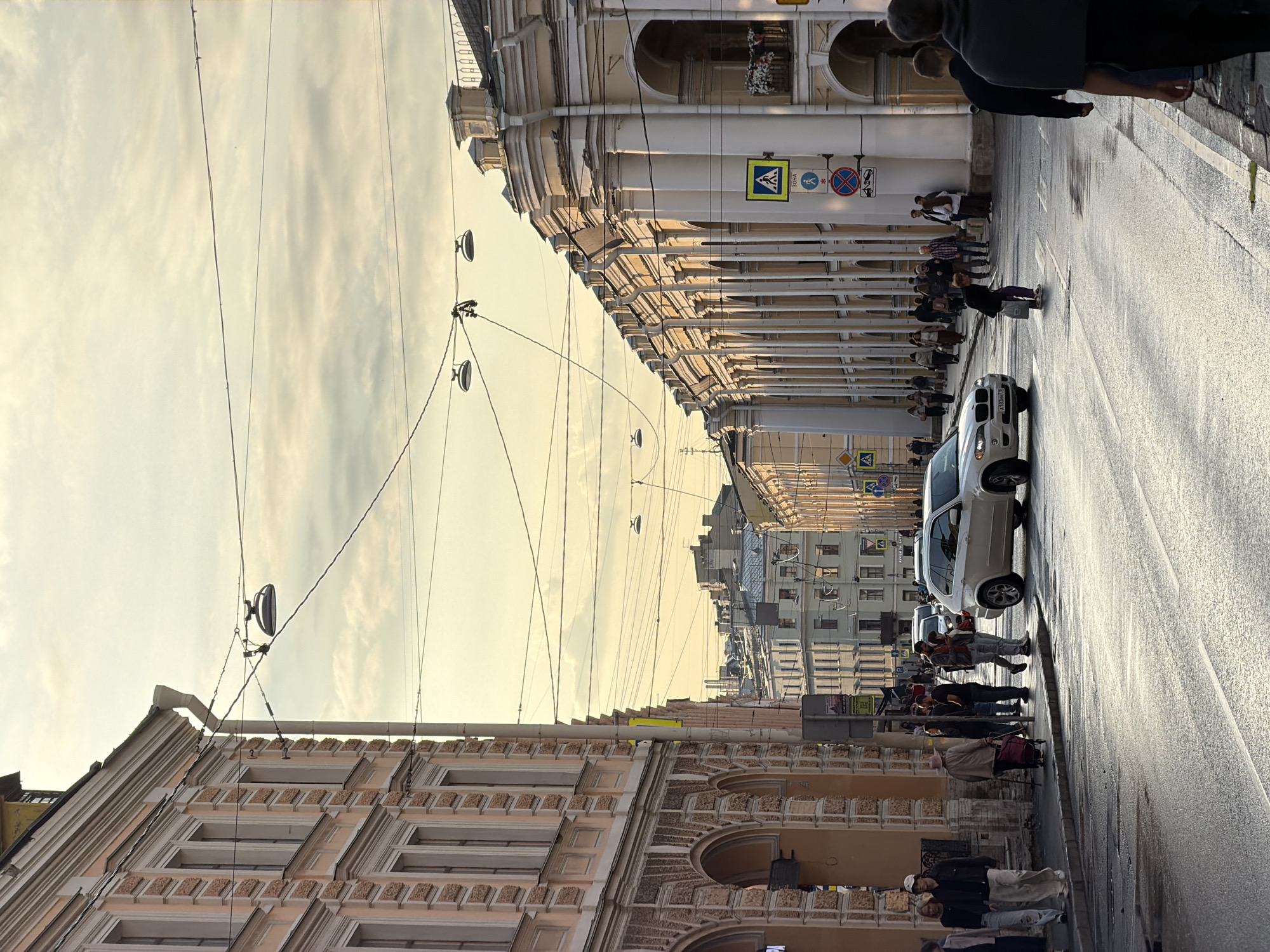 The image shows a street scene in Saint Petersburg, Russia, under a cloudy sky with golden sunlight. The street is wet, suggesting recent rain. On the left, a building with a pale pink facade features arched ground-floor entrances and decorative upper floors. On the right, a long building with a light yellow facade is characterized by a colonnade of large white columns and arched openings. Overhead, numerous electrical wires crisscross the sky, with several round street lamps suspended from them.
Several people are walking on the sidewalk and crossing the street. In the foreground, on the left sidewalk, a group of people are walking away from the camera. On the right sidewalk, more people are visible, some walking, others standing. A white BMW X5 is driving down the street, and another white car is parked further down. Traffic signs are visible, including a pedestrian crossing sign and other regulatory signs. In the background, buildings of various heights and architectural styles line the street, with many windows and rooftop structures. The overall impression is a busy urban street scene in the late afternoon or early evening.