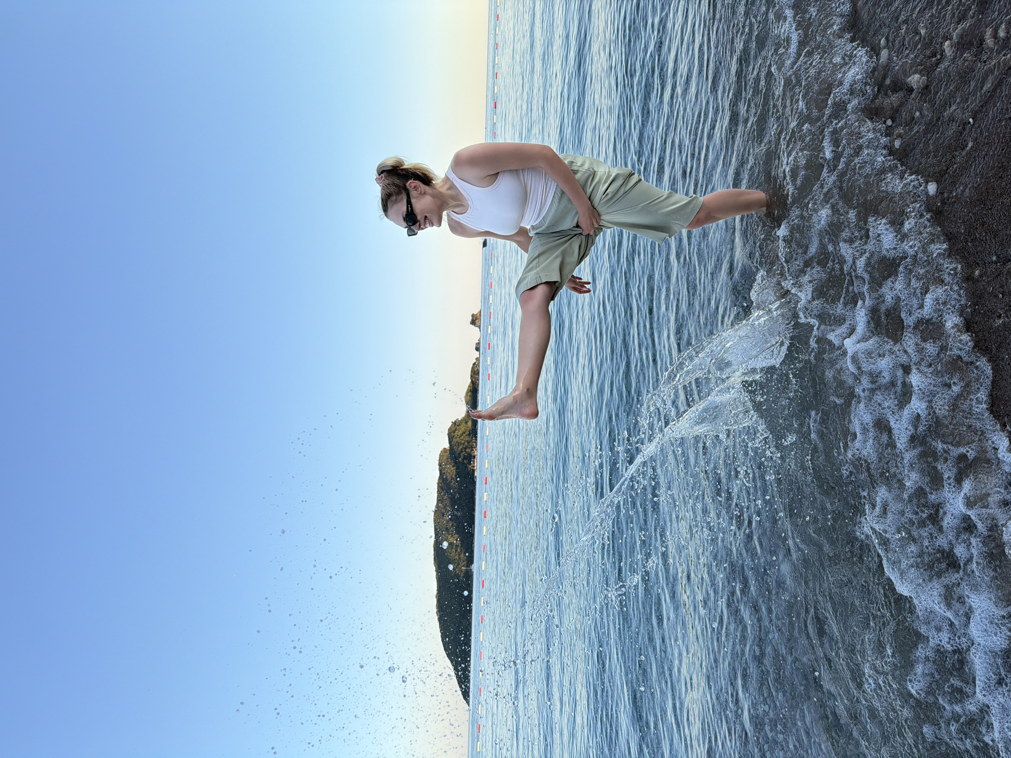 A woman is pictured standing in the shallow water of a beach, kicking her right leg forward and creating a splash. She is wearing a white tank top, light green shorts, and black sunglasses. Her hair is pulled back in a ponytail. In the background, there is a calm sea with a small, green, tree-covered island in the distance. A string of buoys with red and white markers separates the swimming area from the open sea. The sky above is clear and blue. The scene appears to be set in Budva, Montenegro.