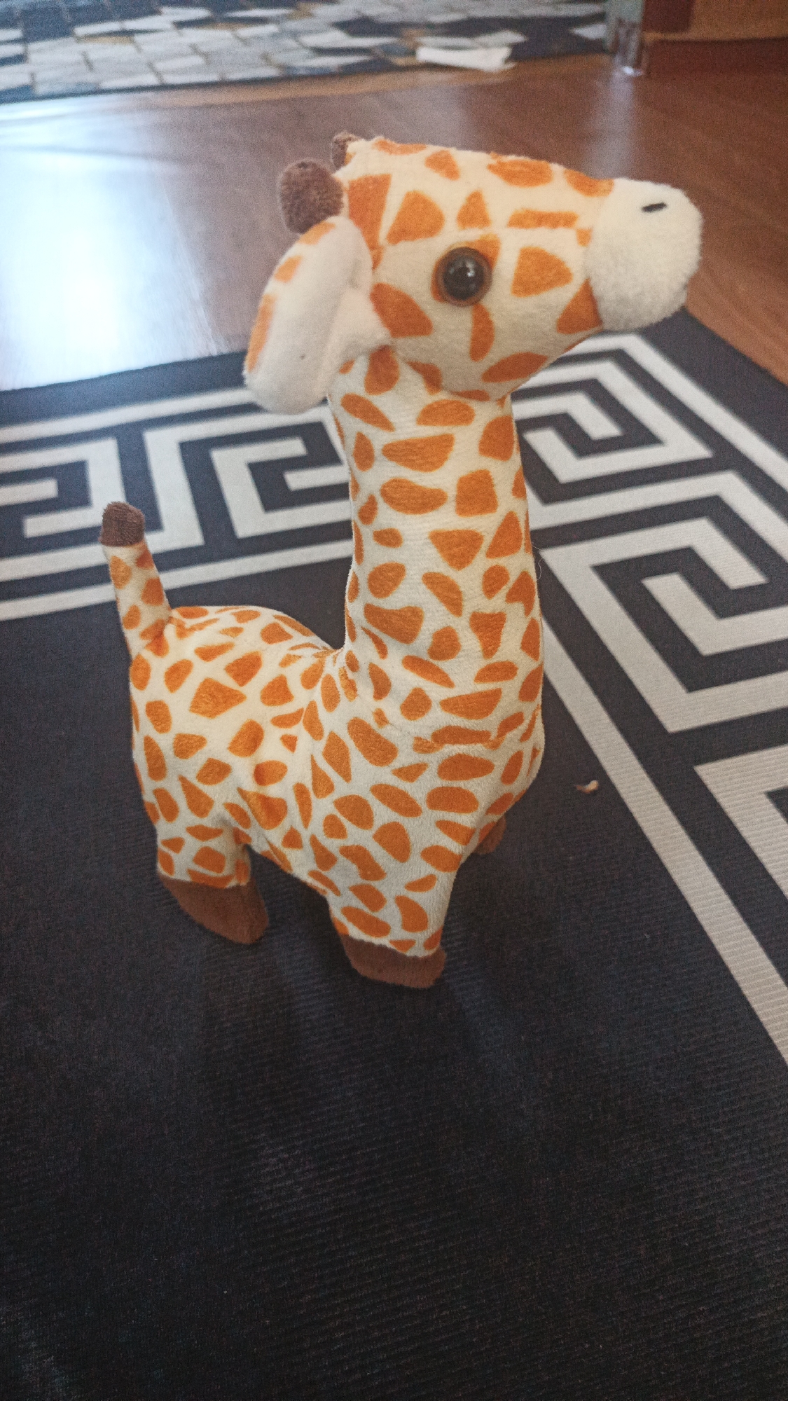 A stuffed giraffe toy stands on a rug. The giraffe is white with orange spots and brown hooves and ears. The rug has a black and white geometric pattern. In the background, a wooden floor and a tiled surface are visible. The scene appears to be indoors, possibly in a residential setting.