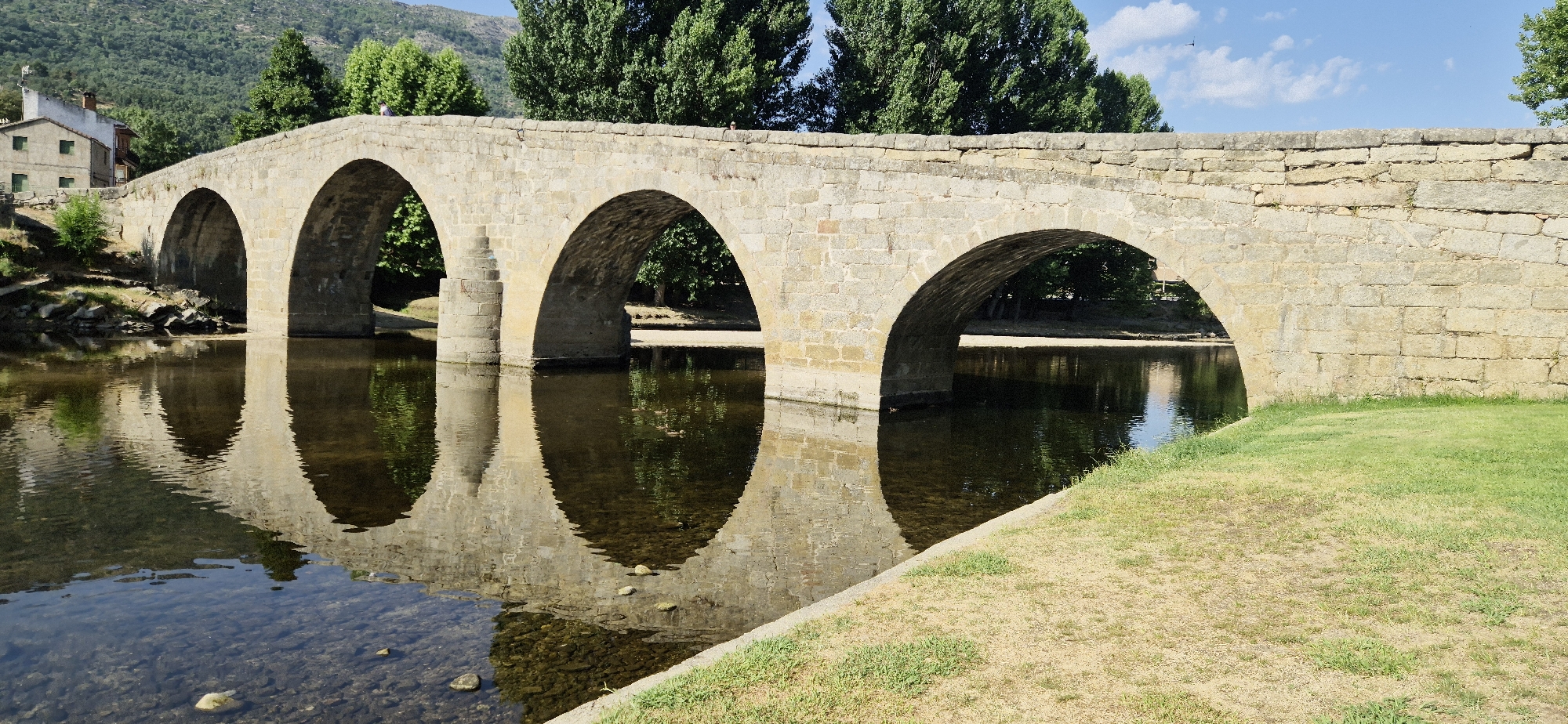 A stone bridge with five arches spans a calm body of water reflecting the structure and the surrounding trees. The bridge is constructed from roughly cut, light-colored stones. The water is clear in some areas, revealing a rocky bottom, and darker in others, showing reflections. On the left bank, a light-colored building with a pitched roof and green-shuttered windows is visible, nestled against a backdrop of trees and a distant, tree-covered mountain. The right bank features a grassy slope leading up to the bridge. Two individuals, appearing as small figures, are standing on the bridge. The sky above is blue with scattered white clouds. The overall scene depicts a tranquil rural landscape.