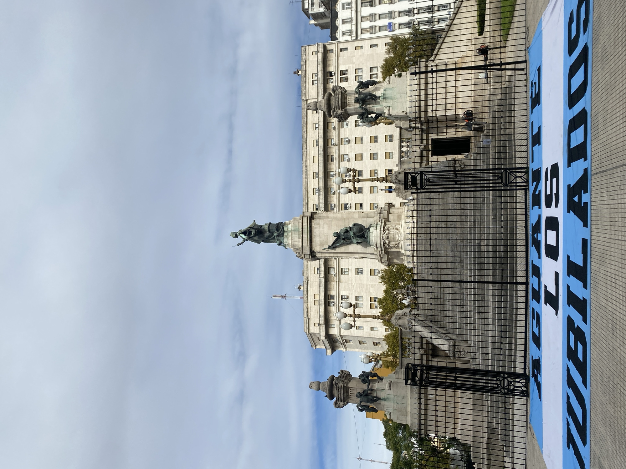 The image captures a view of Plaza de Mayo in Buenos Aires, Argentina. Dominating the foreground is a portion of the monument to General Manuel Belgrano and its surrounding fence. Behind the monument, there's a large government building with multiple floors and windows, typical of the architecture in this historical area. A banner with light blue and white stripes, resembling the Argentinian flag, lies on the ground. Two people, dressed in work clothes are seen, likely tending to maintenance or cleaning tasks around the square. The sky is cloudy, suggesting either an overcast day or early morning.