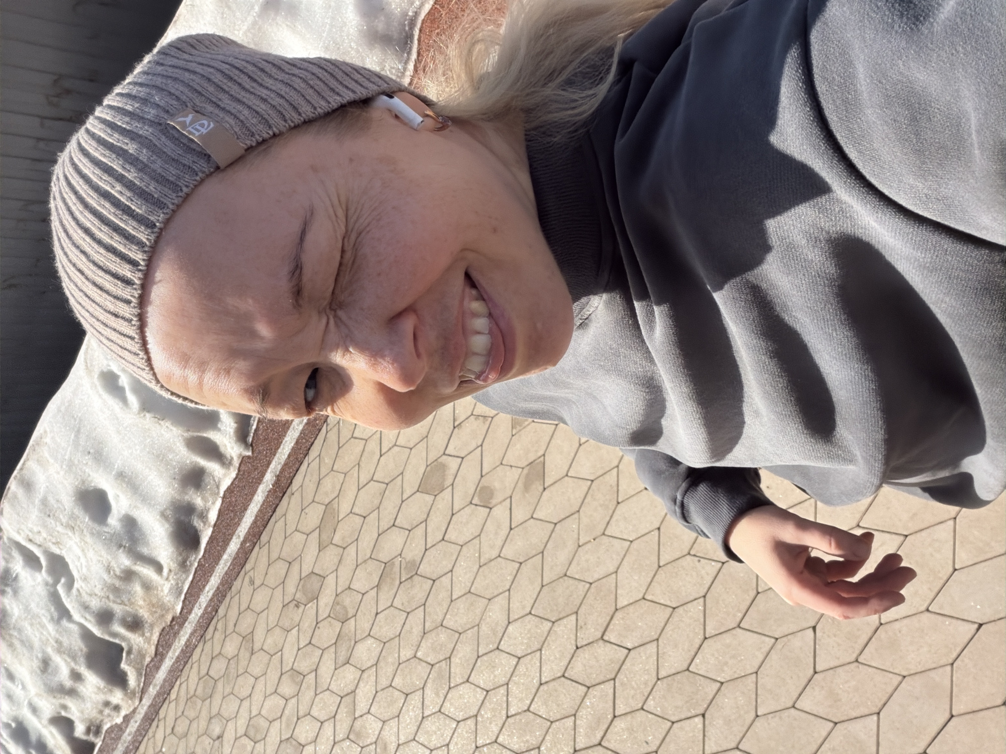 A woman is taking a selfie. She has fair skin, blonde hair, and is wearing a grey beanie and sweatshirt. She is smiling widely, and her eyes are squinted. She is wearing a white earbud in her right ear. To the left of the frame, there is snow and ice, and on the right, the ground is covered in paving stones in a repeating geometric pattern. The location is likely Pervomaysky, Russia.