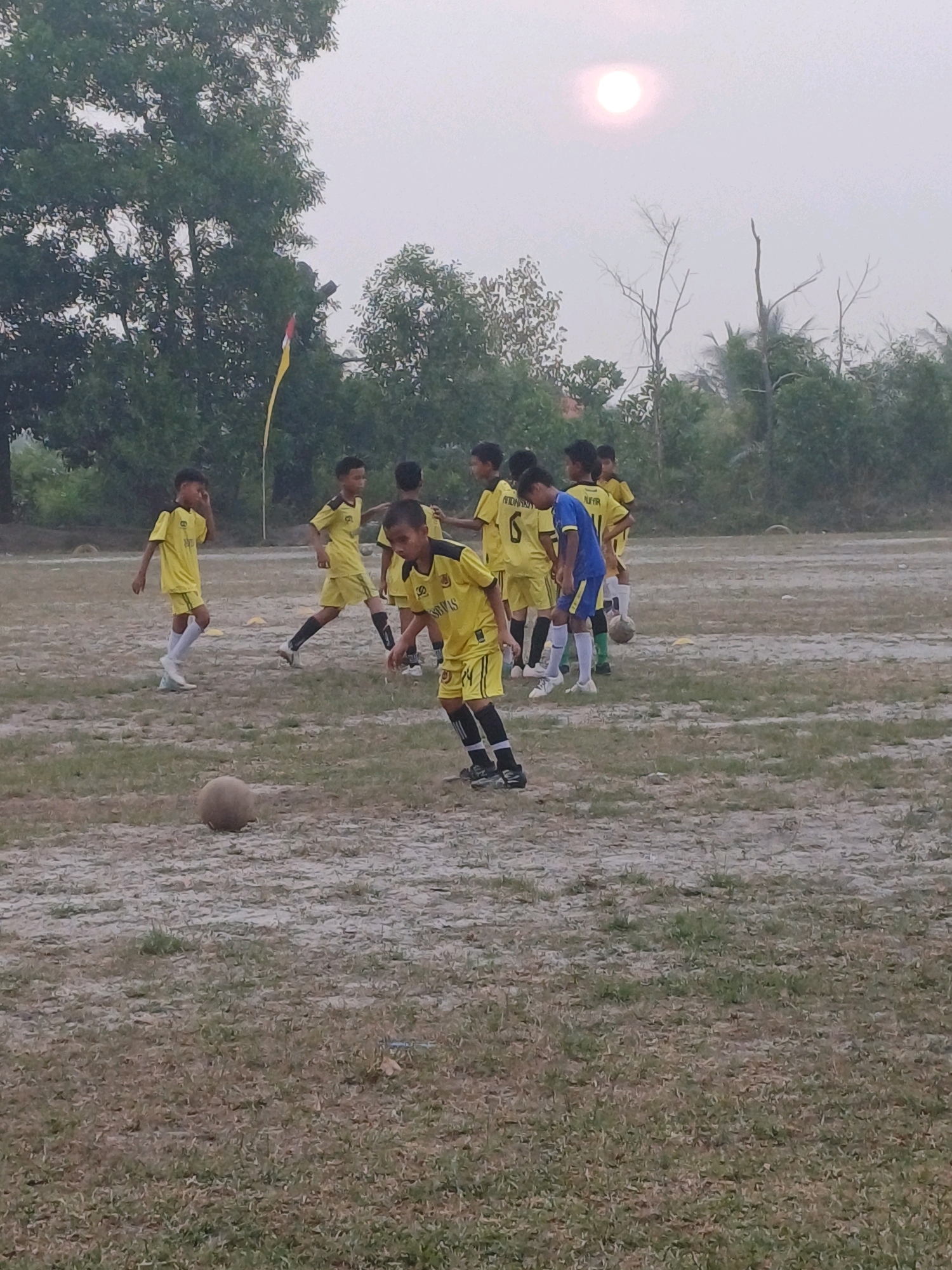 The image depicts a group of young boys in yellow soccer uniforms on a dirt field, likely in Dumai City, Indonesia. The sky is overcast with a pale sun visible in the upper left quadrant. The field is mostly dry and dusty, with sparse patches of grass. Several trees and foliage form a backdrop to the scene. The boys appear to be engaged in a soccer practice or game, with one boy in the foreground focused on a ball in front of him. Another boy in a blue and yellow uniform is visible in the middle ground. The overall impression is an outdoor sports activity in a somewhat rustic environment.