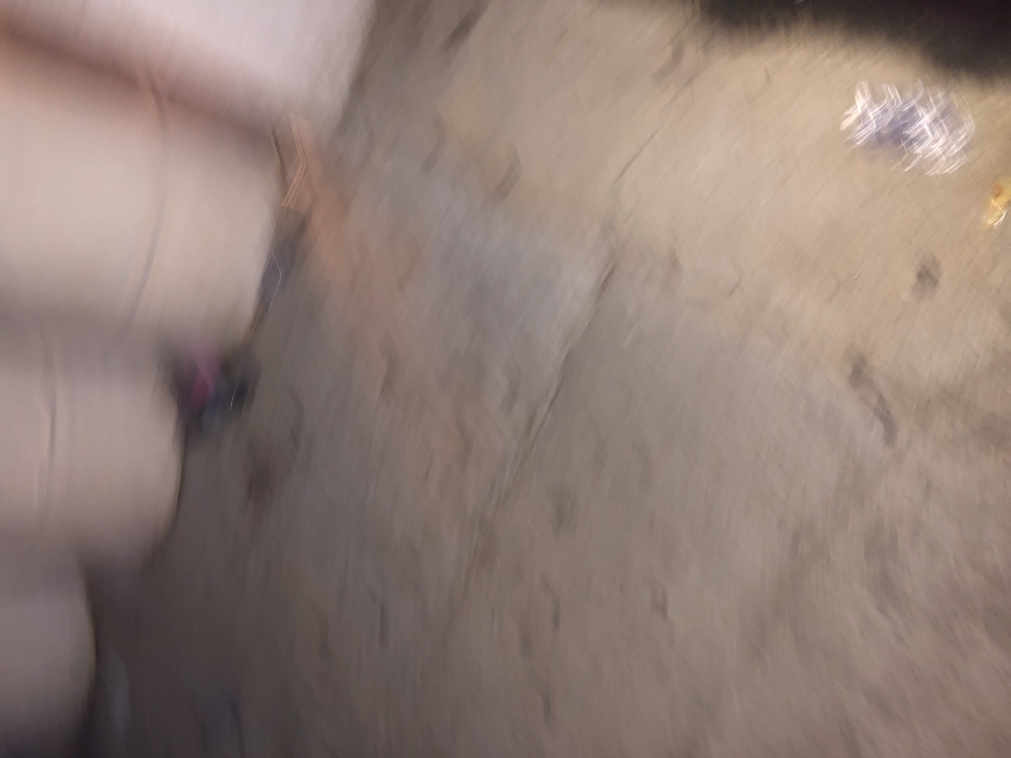 The image is an abstract, motion-blurred depiction of a scene likely on the ground. The main subjects appear to be several stacked, light-colored objects, possibly cushions or soft furnishings, positioned vertically on the left side of the frame. These objects occupy a significant portion of the left and top-left areas.
The ground surface, taking up the majority of the image, is textured and appears to be made of concrete or a similar material, with variations in tone suggesting dirt or stains. In the upper right quadrant, a blurred, shiny object, possibly a crumpled piece of plastic or a collection of small items, is visible. Another small, yellow object is also present near this cluster. A small, dark, and blurry object, possibly a shoe or a bag, is visible between the stacked objects on the left.
The overall impression is one of a casual, indoor or outdoor setting where items have been placed or discarded on the floor. The motion blur suggests the image was taken while the camera was moving or that the subject itself was in motion. No specific time of day or weather conditions can be inferred. No text is visible in the image. The context of Howayi, Nigeria, provides a geographical location but does not offer specific visual cues within the image itself.