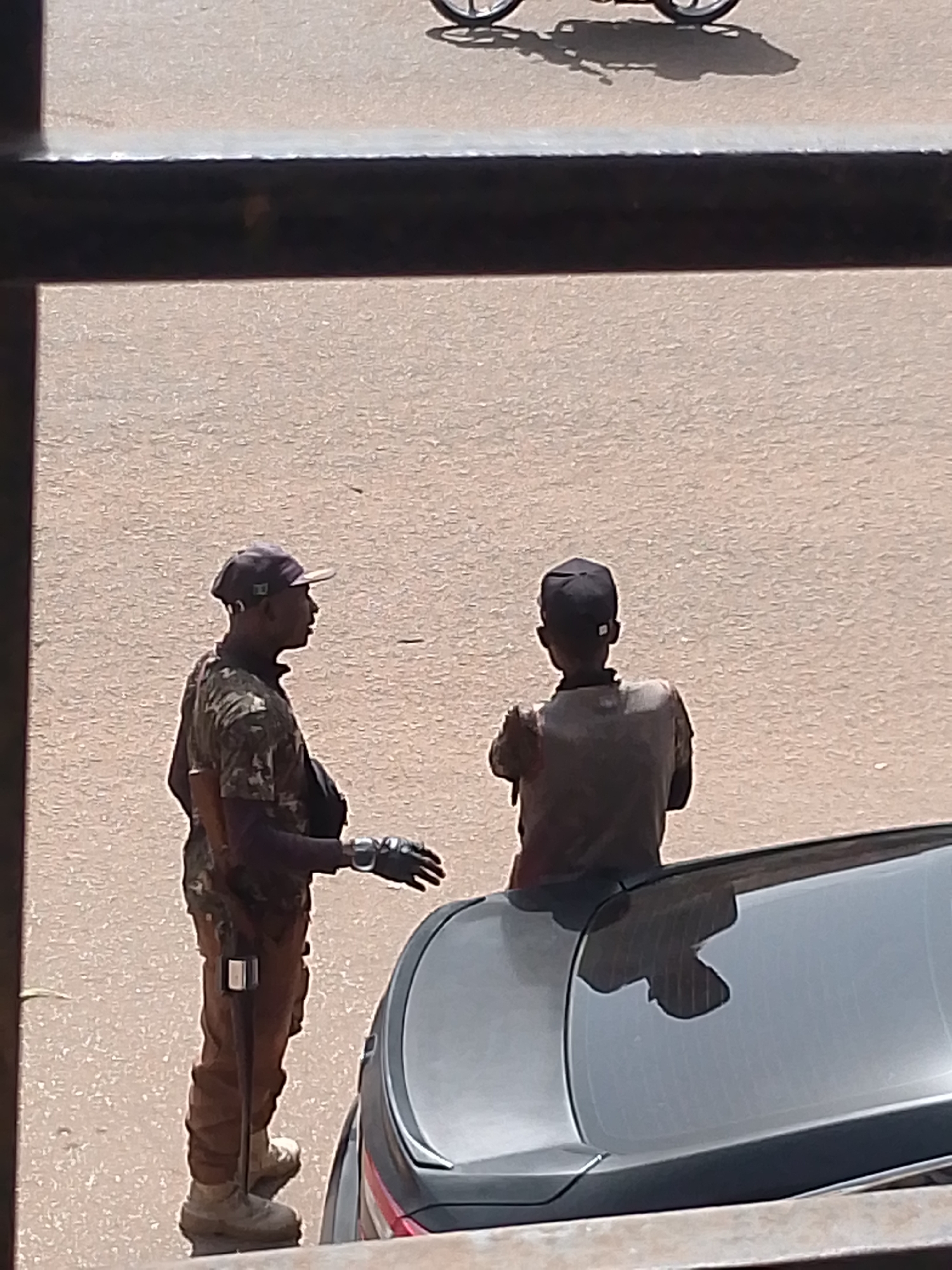 In Bukuru, Nigeria, two individuals stand on a paved surface. The individual on the left is facing right, wearing a camouflage patterned shirt, brown pants, and a cap. They have a rifle slung across their body, with a black glove on their right hand. The individual on the right is facing away from the camera, wearing a dark shirt and a black cap. They are positioned next to a dark-colored car. The background shows a blurred, tan-colored ground and a hint of a bicycle in the upper portion of the image. A dark horizontal bar is visible in the foreground, obscuring the lower part of the scene.