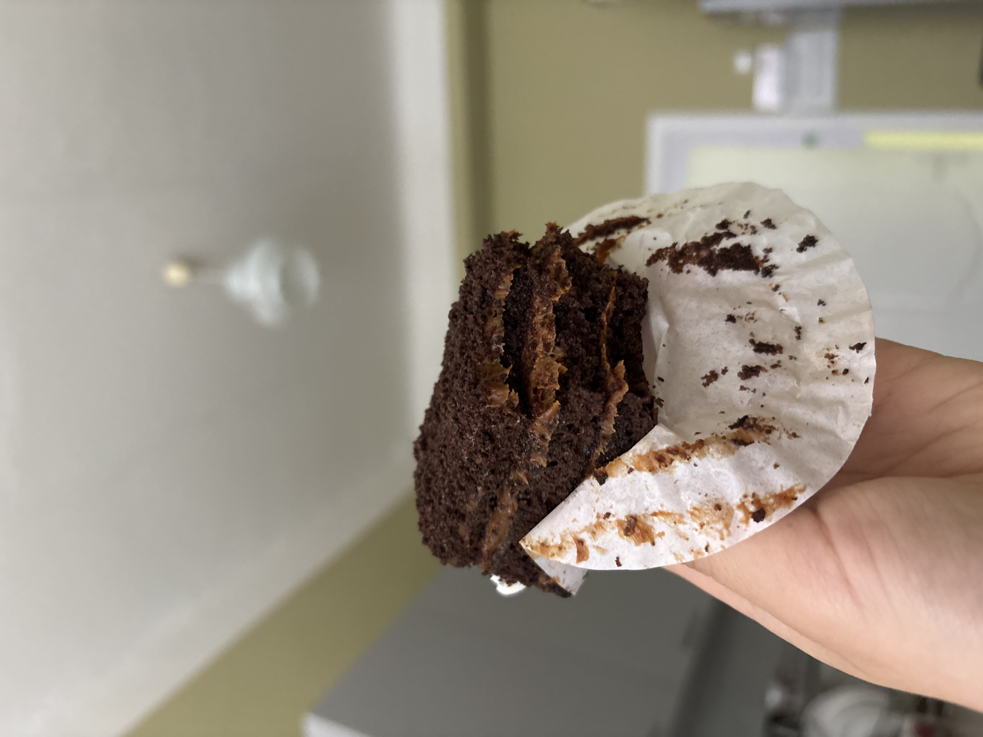 A close-up shot shows a partially eaten chocolate cupcake held in a hand. The cupcake is dark brown, with some frosting visible within its layers. The cupcake paper is white and stained with chocolate residue. The background is blurred but suggests an indoor setting with a light fixture on the ceiling and a white surface, possibly a cabinet or refrigerator, behind the hand. It is not possible to determine the location as Chaikovsky, Russia, based on this information.