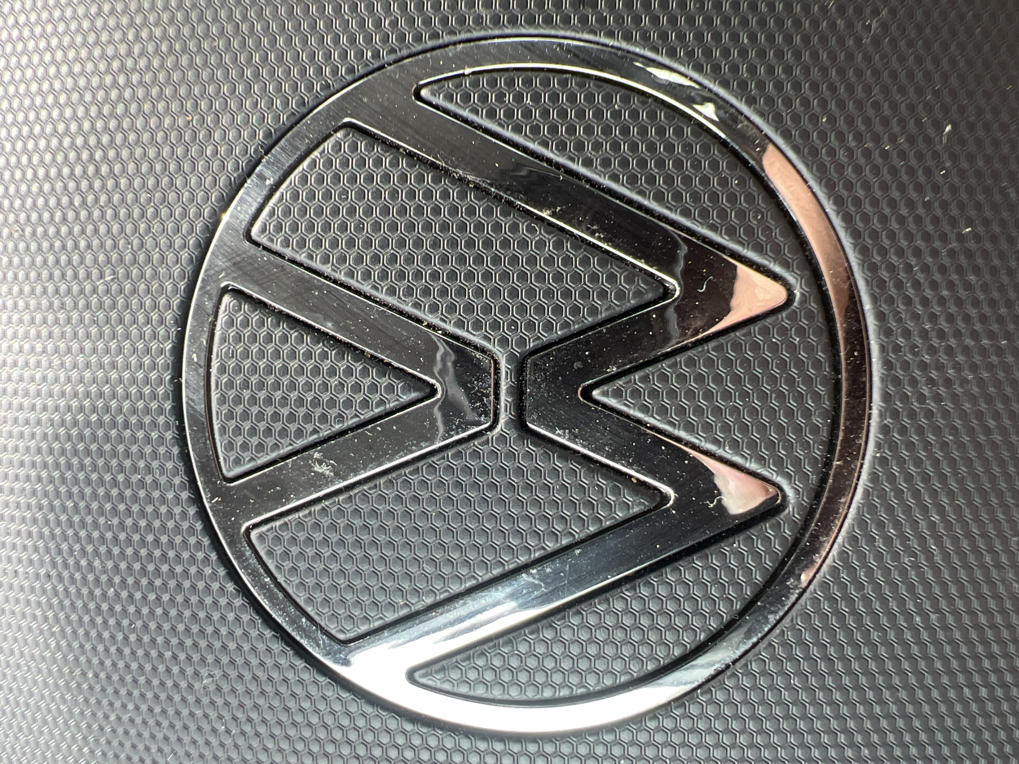 The image shows a close-up, detailed view of the Volkswagen logo. The logo is circular with a metallic finish and is embossed on a textured black surface that resembles a honeycomb pattern. The logo itself is a stylized "VW" within the circle, with the "V" positioned above the "W". Reflections are visible on the metallic surface of the logo, indicating it is likely a car emblem or part of a car's interior. The background texture is consistent and appears to be a durable material. There are no people or other objects visible in the frame. The lighting suggests an indoor environment, possibly artificial light. The image's focus is sharp on the logo and the immediate surrounding textured surface. The location in Siegen, Germany is not discernible from the visual information provided in the image.