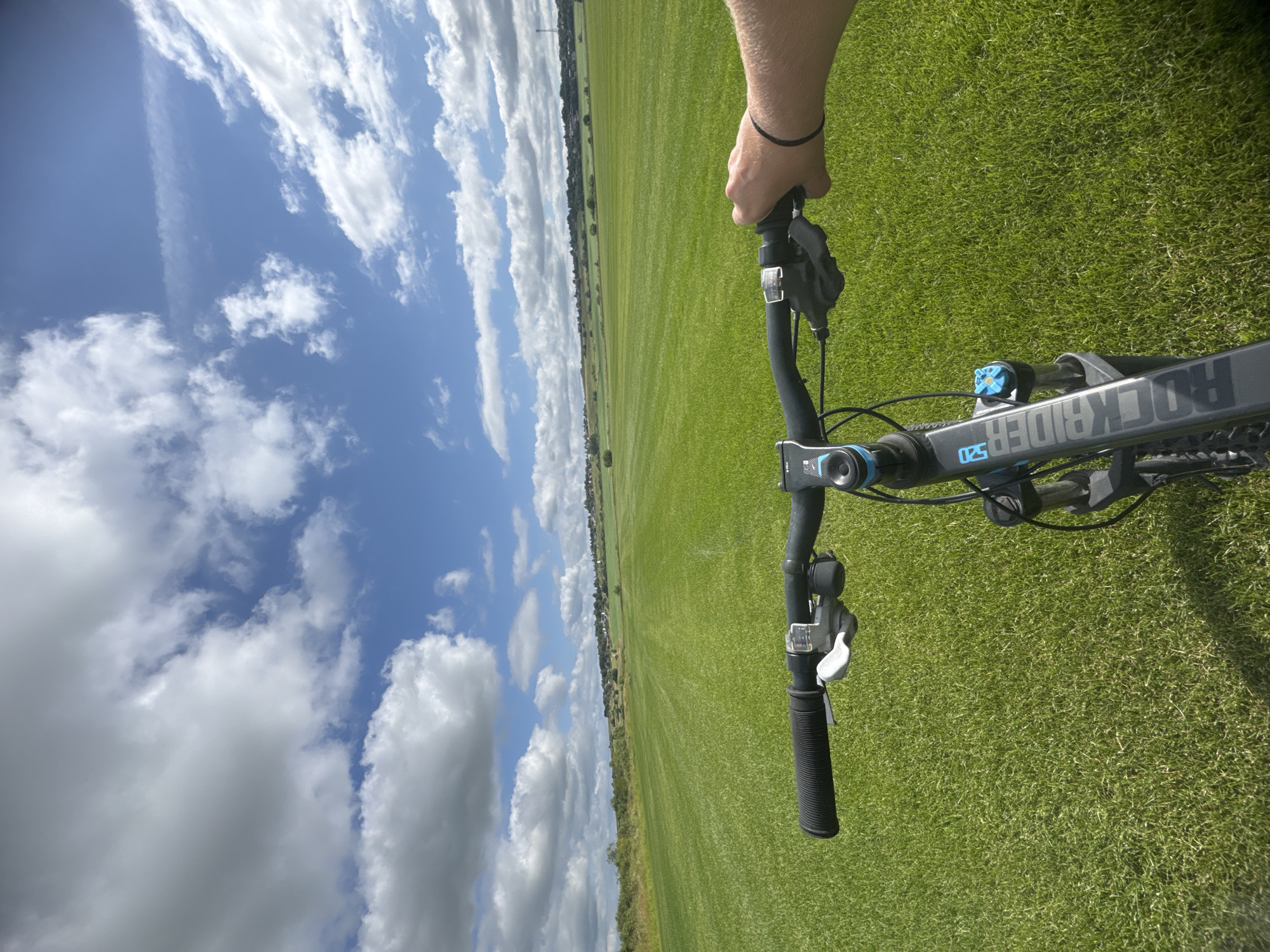 The image is a first-person perspective shot from a bicycle's handlebars. The bicycle has a black frame with "ROCKRIDER 520" printed in blue and white lettering. The handlebars are black, with a white brake lever and a bell. A person's right hand, wearing a black bracelet, is gripping the right handlebar. The bicycle is positioned on a vast, well-maintained green lawn. In the background, the lawn extends to a gently rolling landscape with scattered trees and a distant line of houses. Above, a bright blue sky is filled with large, white cumulus clouds. The image appears to depict an outdoor recreational area, potentially a park or open field, in Kempelevo, Russia.