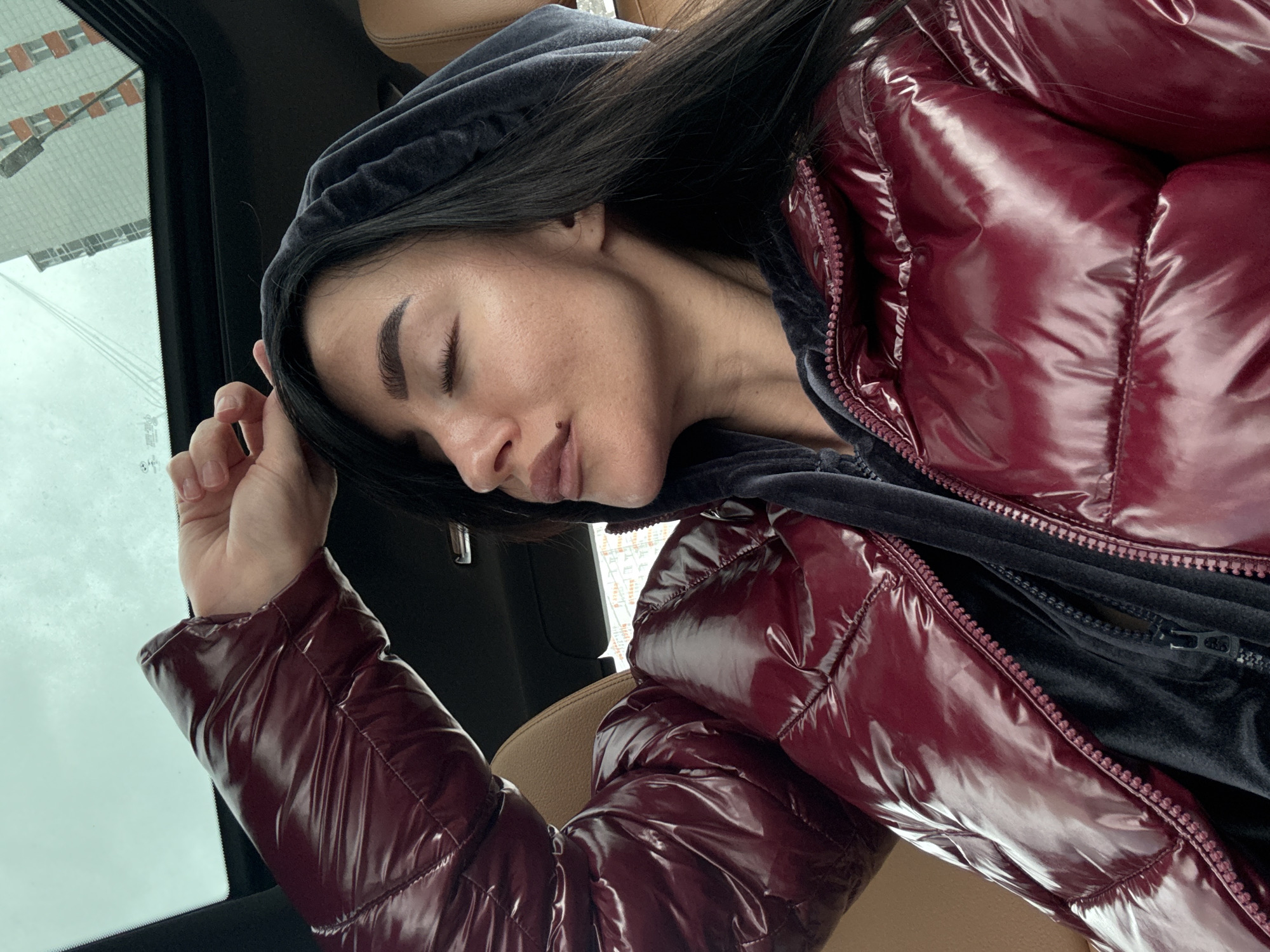 A woman is seated in a car wearing a maroon puffy jacket over a hooded velvet sweatshirt. The woman has dark hair, closed eyes, and is resting her head on her hand. Outside the car window, there are buildings with construction cranes visible. It's daytime and appears overcast outside. Based on the presence of specific architectural elements and construction cranes, the location is plausibly in the city of Krasnoyarsk, Russia.