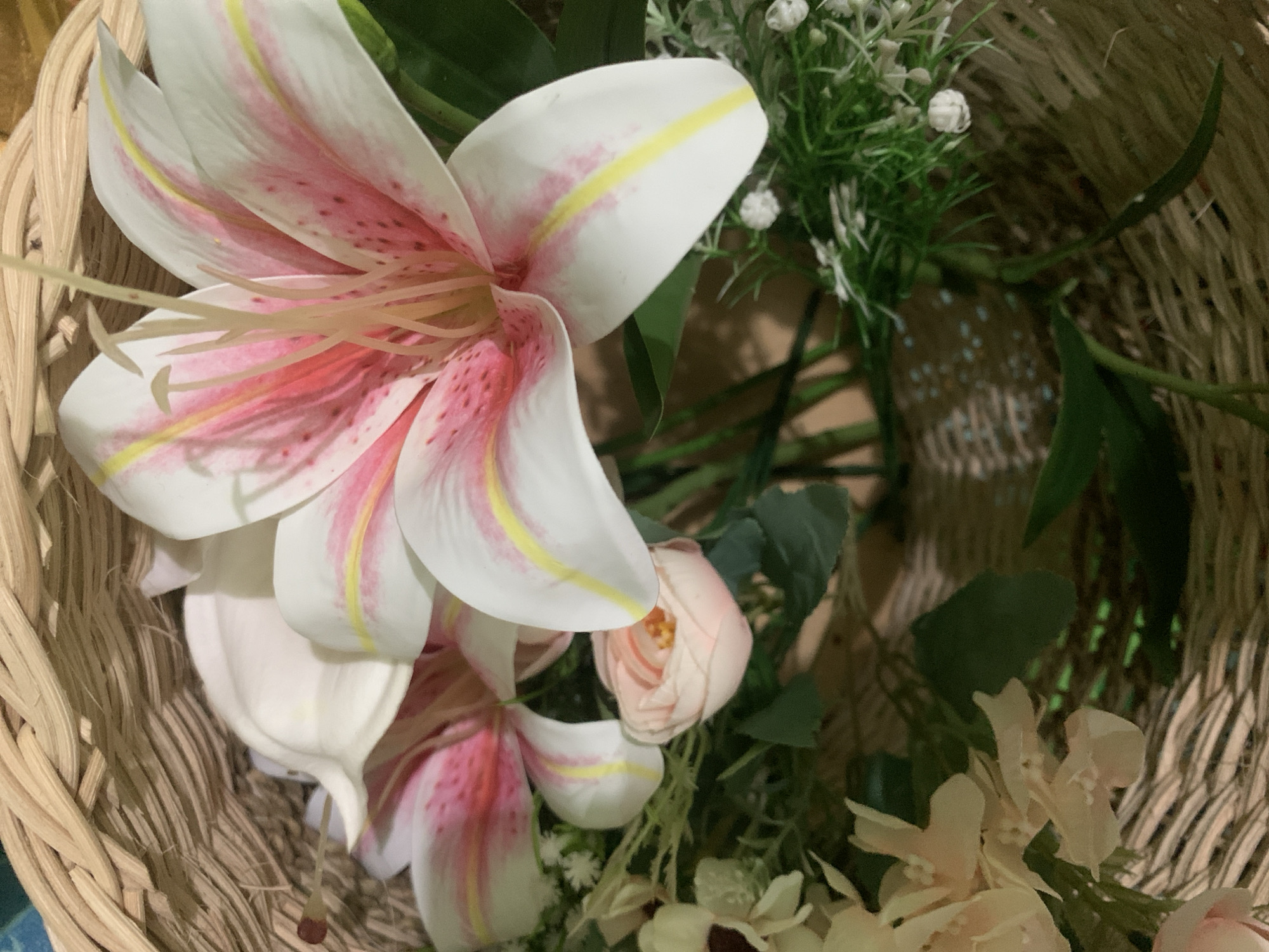 The image shows a close-up view of a woven basket filled with artificial flowers. The flowers are predominantly white with pink and yellow accents, resembling lilies. There are also smaller pink roses and clusters of small white flowers. Green artificial foliage is interspersed among the flowers. The basket appears to be made of light brown wicker. The lighting is soft and diffused, with some shadows visible within the basket. There are no people or animals present in the image. The scene is a still life arrangement of decorative flowers. The provided context indicates the location is Kuala Pembuang, Indonesia, however, this information cannot be visually corroborated from the image itself.