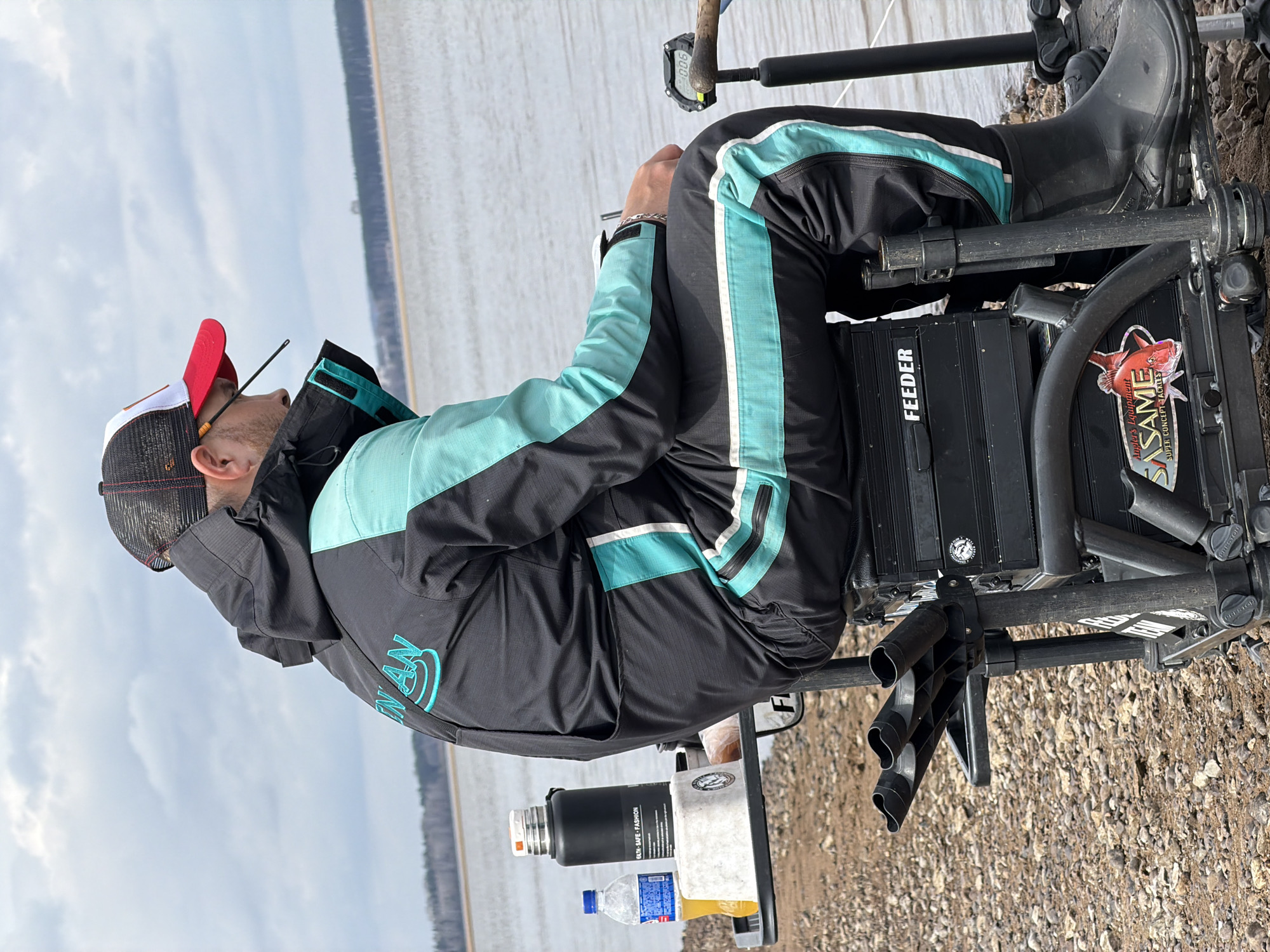 A man wearing a black and red cap and a black jacket with light blue accents is sitting on a black fishing chair beside a body of water. He is facing away from the camera, and the background shows a lake or river under a cloudy sky. There are items next to him on a platform connected to the chair, including a thermos, bottles, and other fishing-related accessories. The ground is gravelly. The name "Chaykovsky" is not identifiable in the image but is likely the location as stated in the prompt.