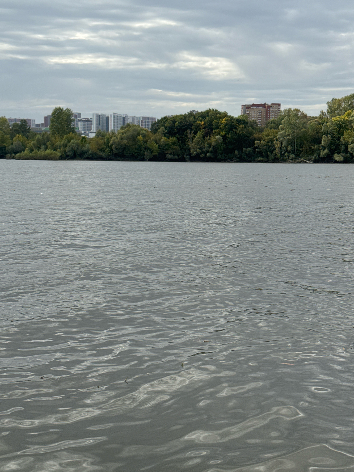 A wide body of water, likely the Ufa River, occupies the foreground of the image. The water's surface is rippled, with sunlight glinting off the crests of small waves. In the middle ground, a dense line of trees with green and yellow foliage borders the water. Behind the trees, a cityscape is visible. Several modern, multi-story buildings, predominantly in shades of white, grey, and beige, rise above the treeline on the left side of the frame. To the right of these buildings, a reddish-brown apartment building with visible windows stands out. The sky above is overcast with a heavy layer of grey clouds, obscuring any clear view of the sun. The overall impression is that of an urban waterfront on a cloudy day, possibly in the city of Ufa, Russia, given the architectural style and vegetation. There are no people or animals visible in the frame, and no discernible actions or interactions are taking place.